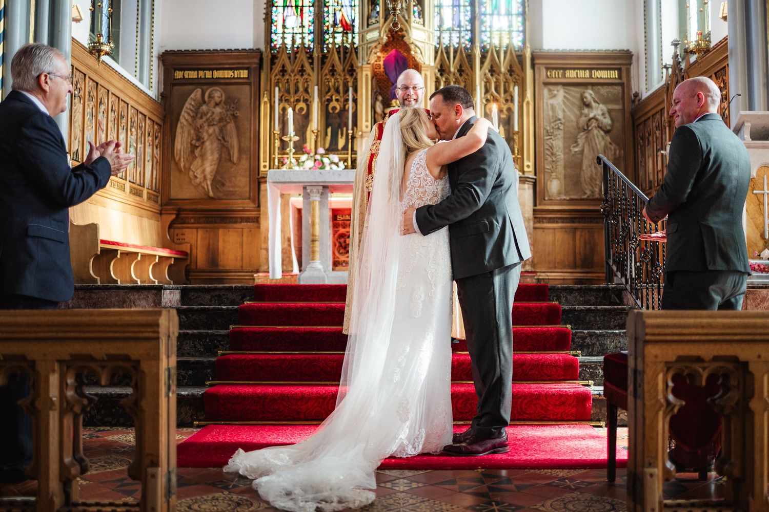 Bride and groom share first kiss at church altar with red carpet steps and ornate decor during wedding ceremony.