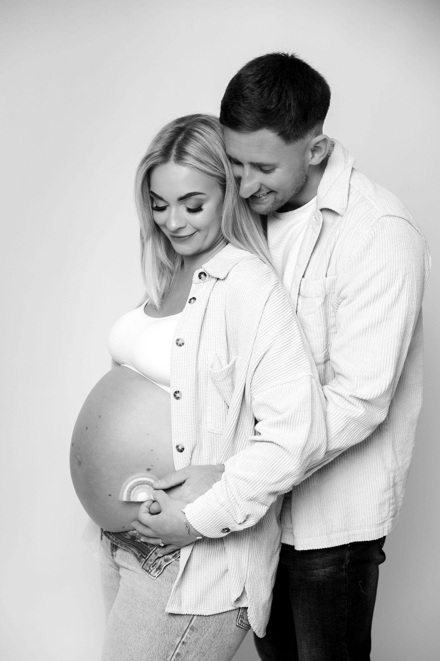 Black and white photo of a smiling pregnant couple embracing, gently cradling the baby bump together.