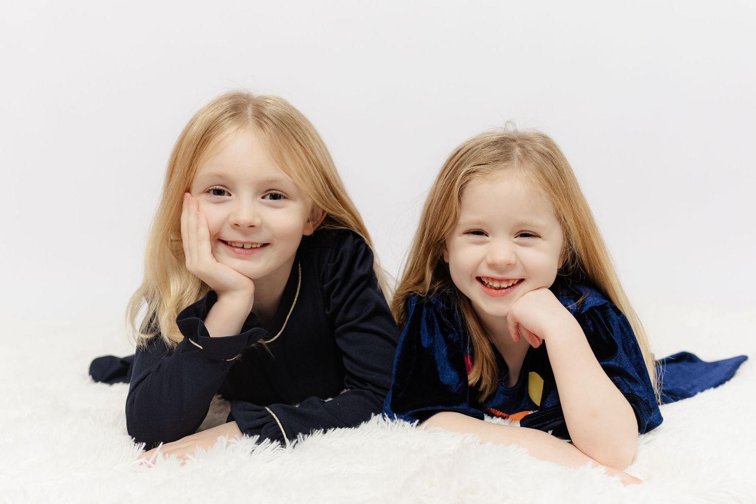Two smiling blonde girls in velvet pajamas lying on a fluffy white rug in a studio portrait photo.