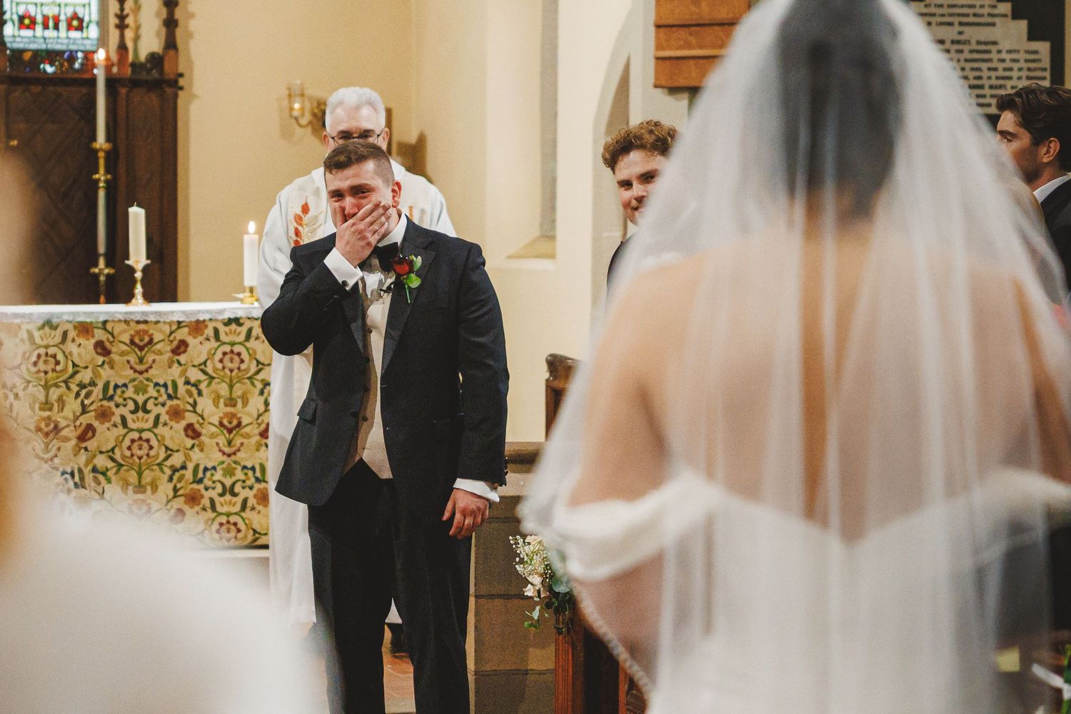 Emotional groom covering his mouth in tears as bride walks down the aisle in a church wedding ceremony.