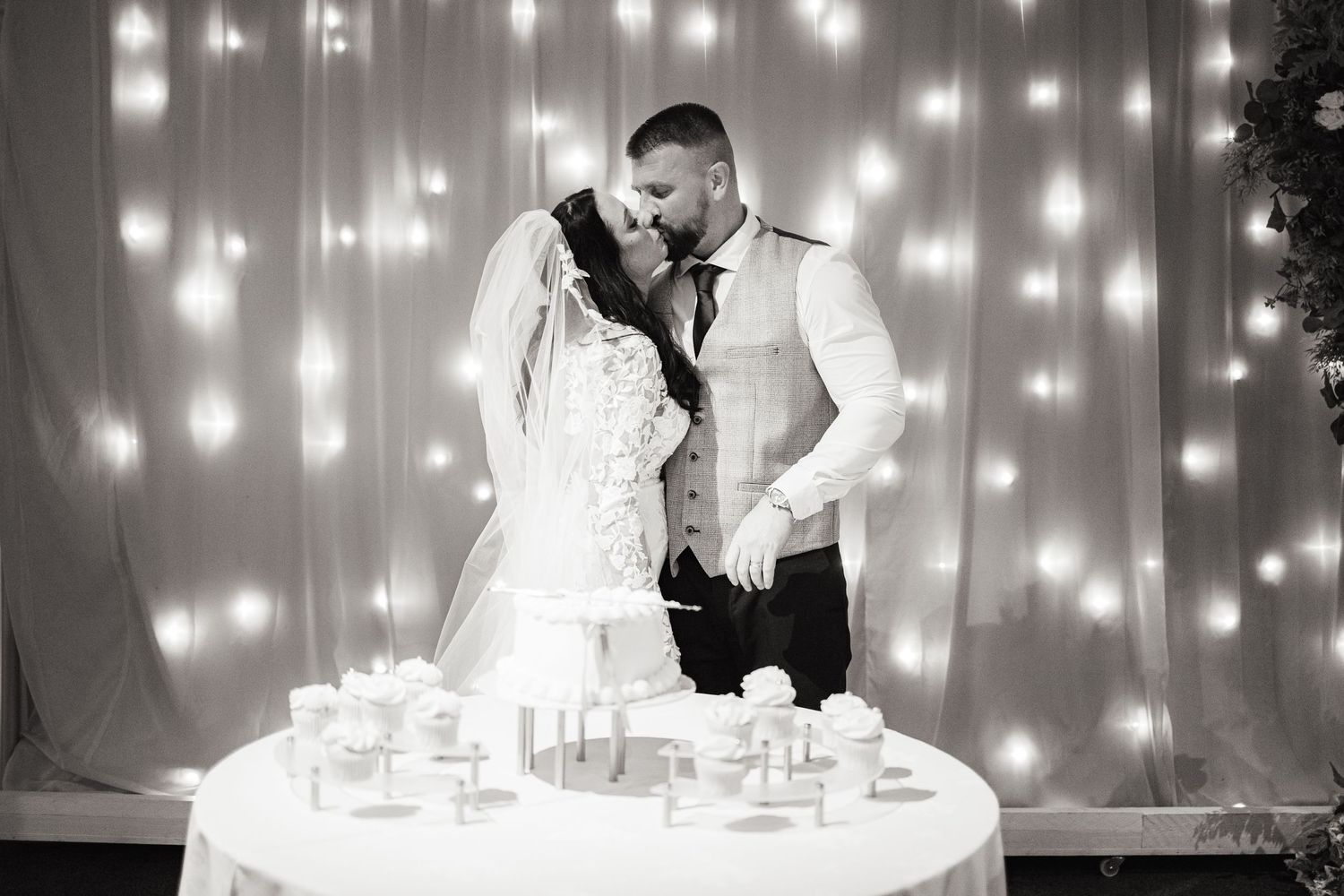 Black and white photo of a bride and groom kissing while cutting their wedding cake, with fairy lights backdrop.