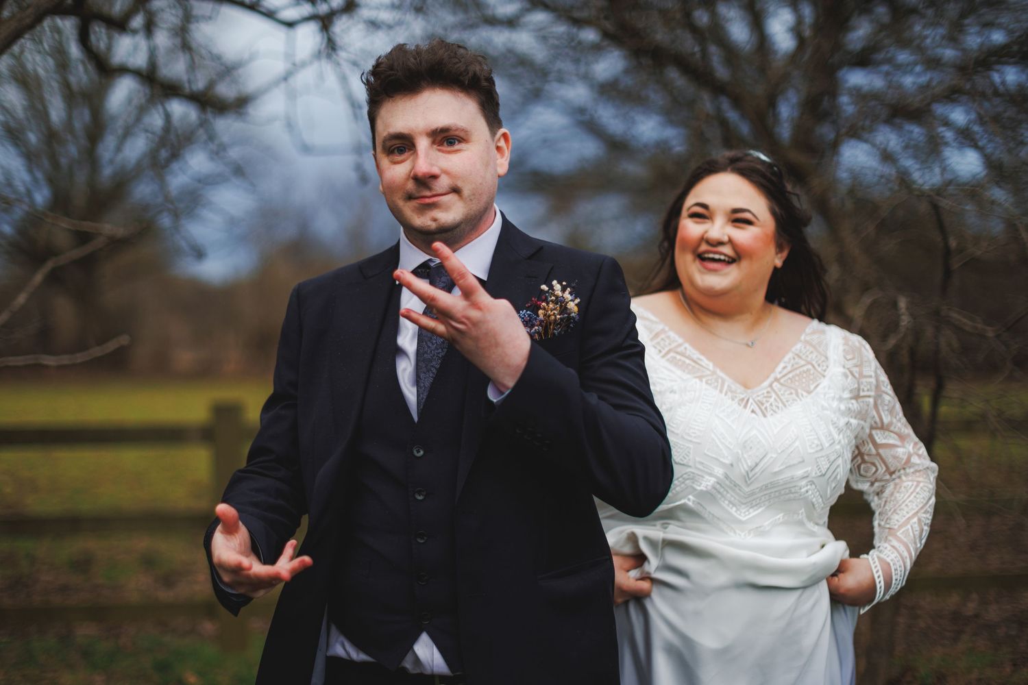 Bride and groom laughing outdoors at rustic wedding venue, groom in navy suit, bride in white lace dress.