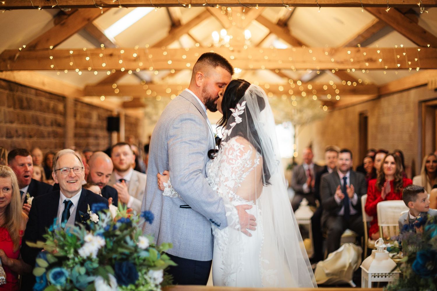 Bride and groom share first kiss at rustic barn wedding ceremony with fairy lights and guests applauding in background.
