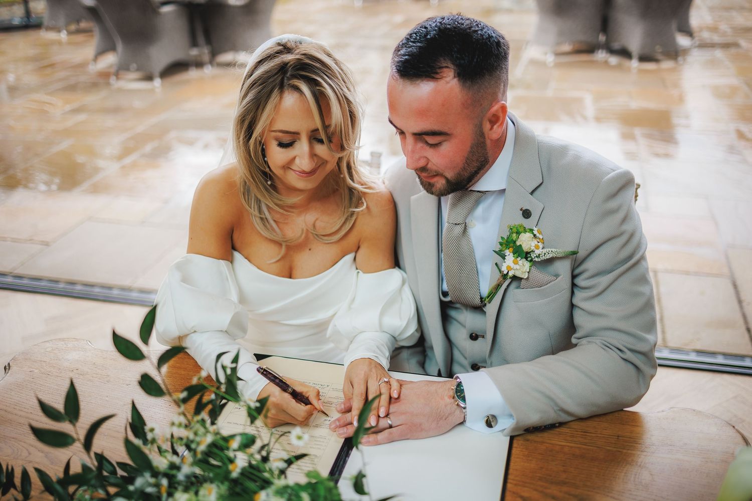 Bride and groom signing wedding register together, bride in off-shoulder gown, groom in grey suit with boutonniere.