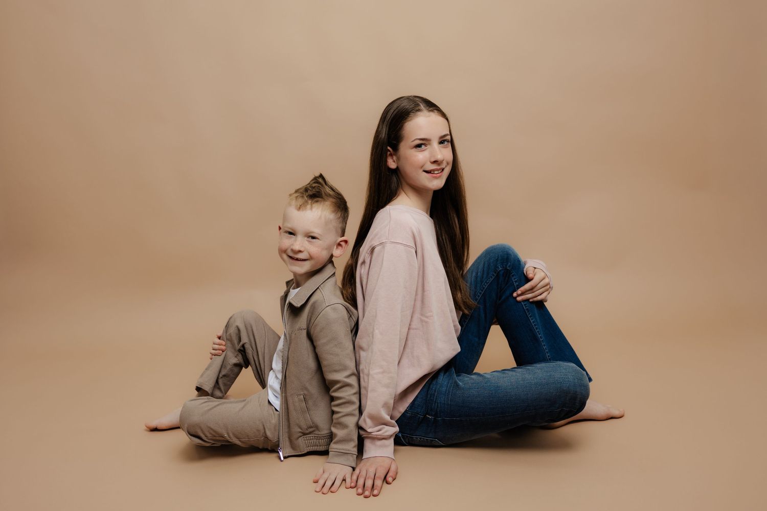 Two children, a boy and girl, sit back-to-back smiling on a beige studio backdrop in casual stylish outfits.