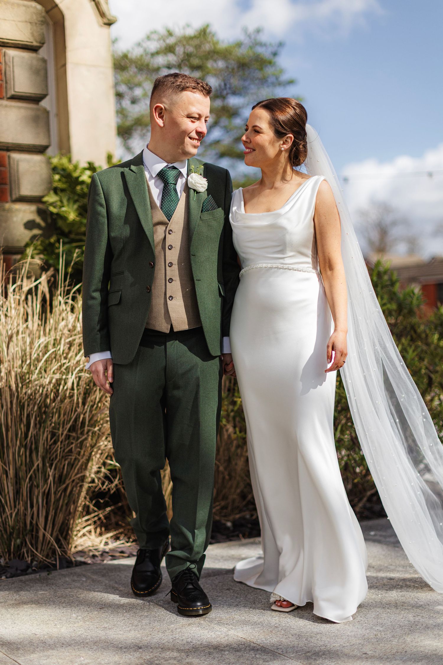 A bride and groom walk together outdoors, she in a white gown with veil, he in a green suit with beige waistcoat.