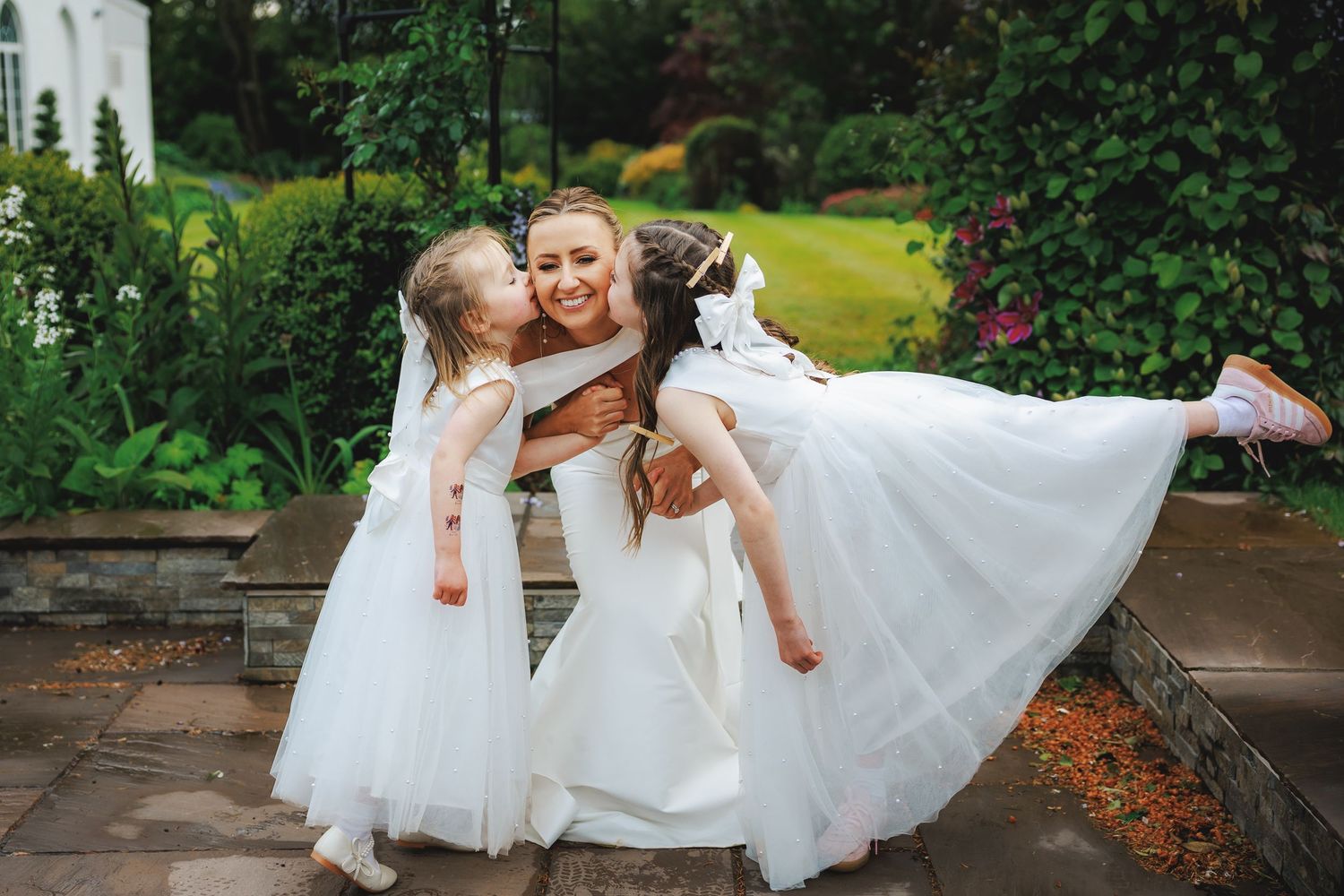 Bride in white gown hugged by two flower girls in white dresses in a lush garden setting.