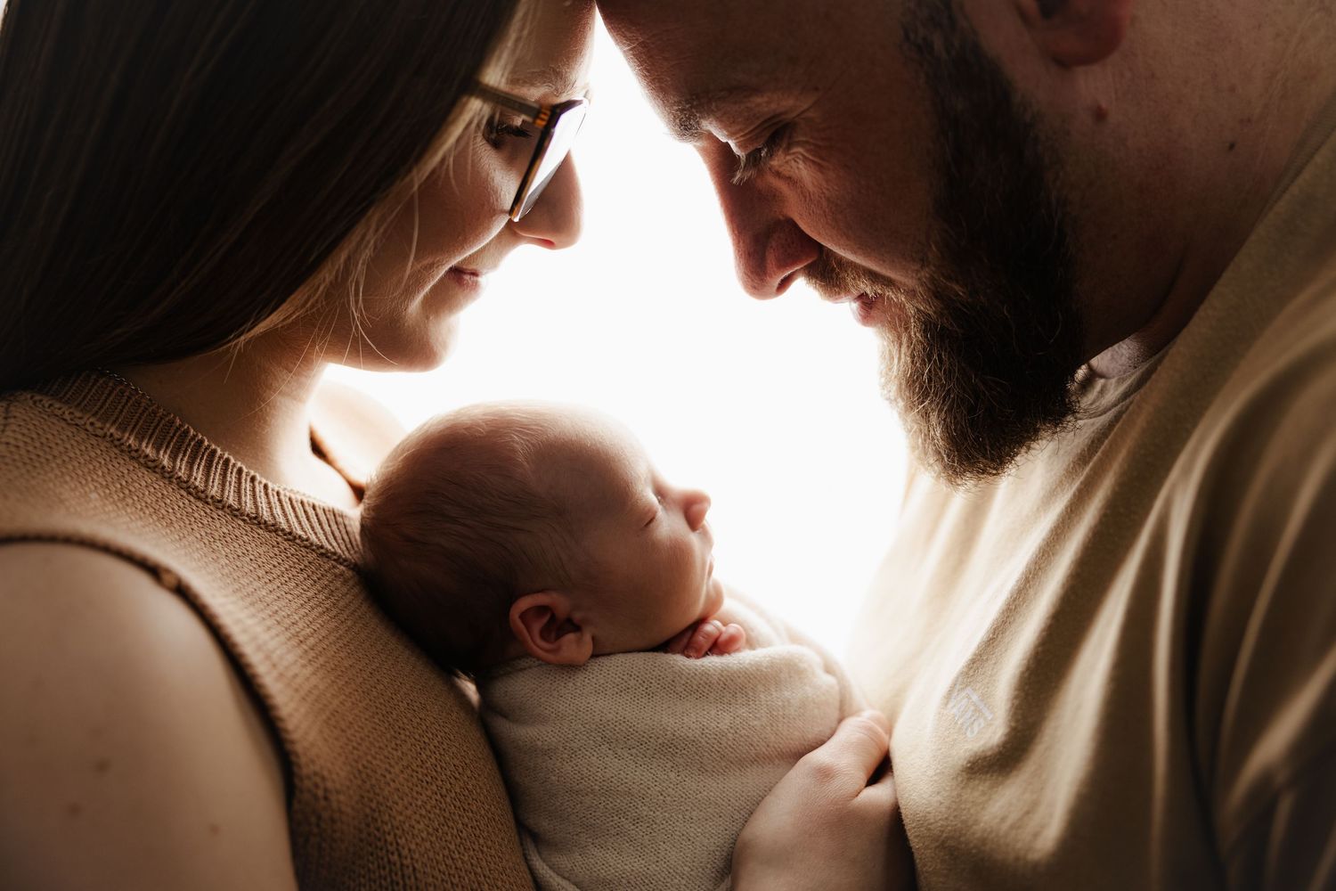 A couple with a beard and glasses tenderly hold their newborn baby, sharing an intimate family moment in soft backlit light.