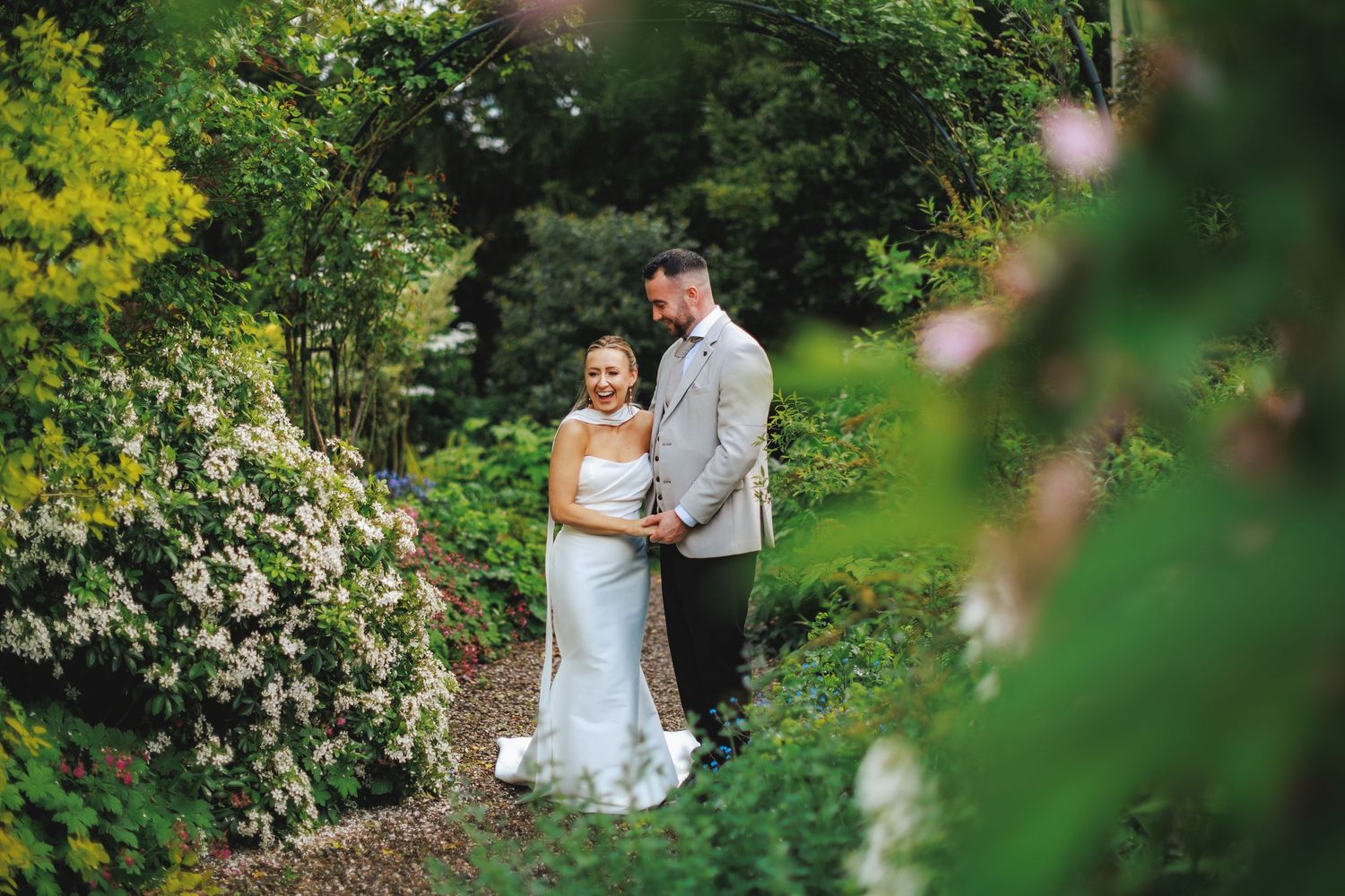 A bride and groom embrace in a lush garden path surrounded by blooming flowers and greenery on their wedding day.
