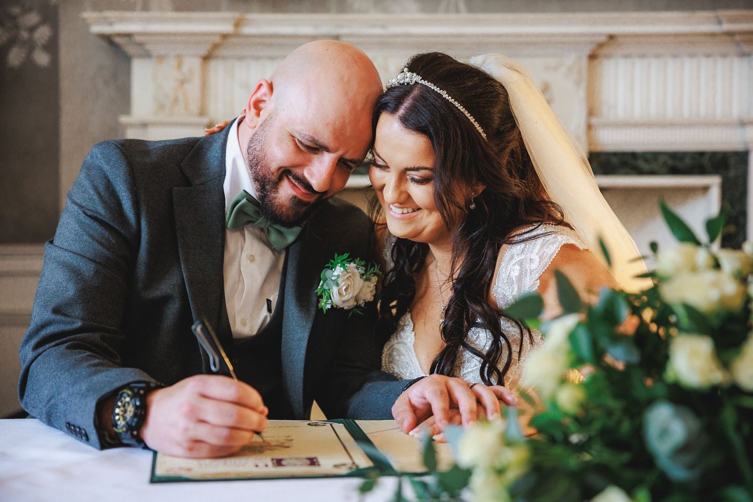 A smiling bride and groom signing their wedding register together at a ceremony table with floral decorations.
