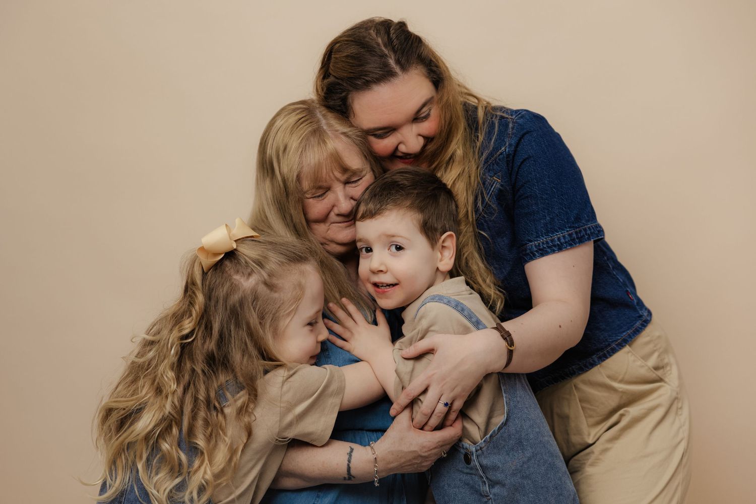 Family portrait of two adults hugging two young children against a beige background in a warm studio photo.