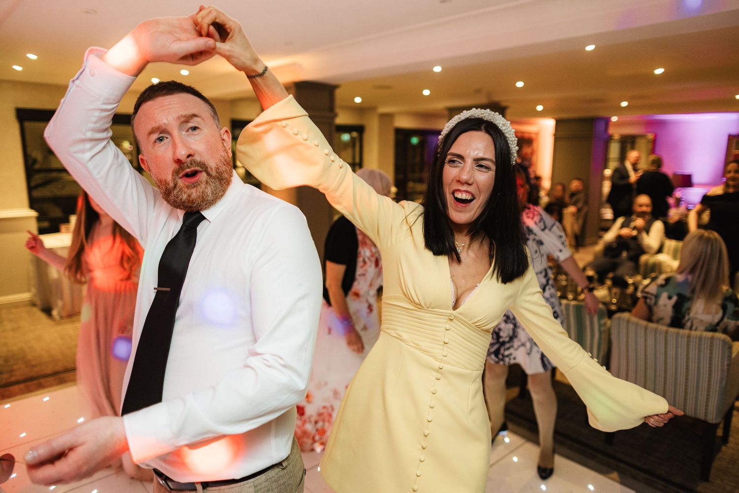 Couple dancing joyfully at a wedding reception, woman in yellow dress with tiara, man in white shirt and black tie.