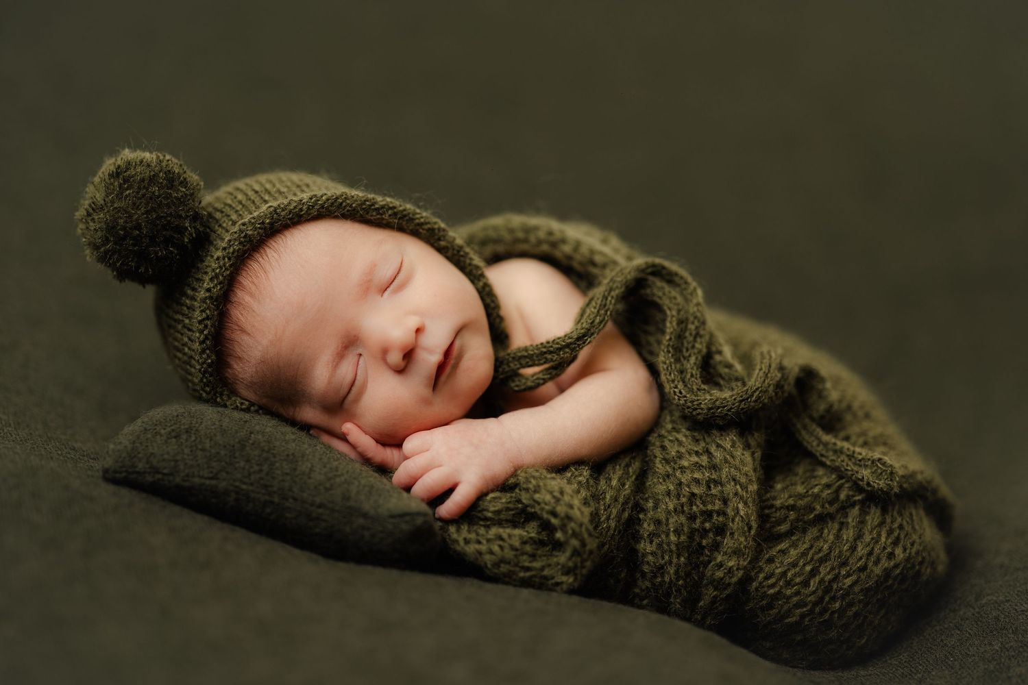 Sleeping newborn wrapped in olive green knit swaddle and hat on dark background.