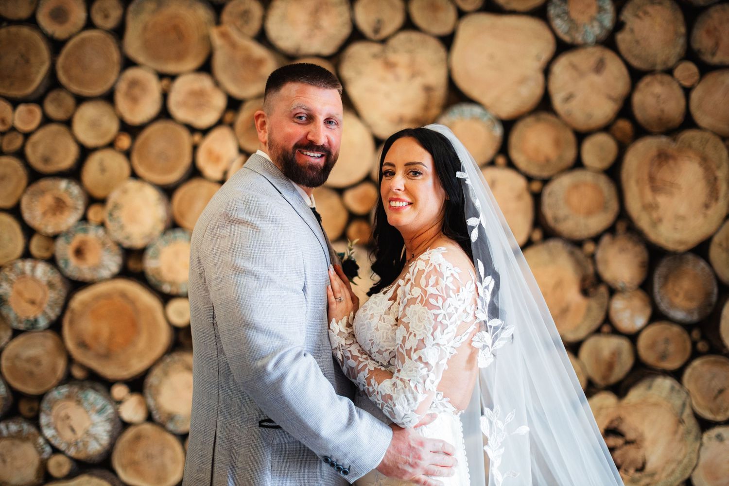 A smiling bride and groom pose together in front of a rustic log wall backdrop on their wedding day.