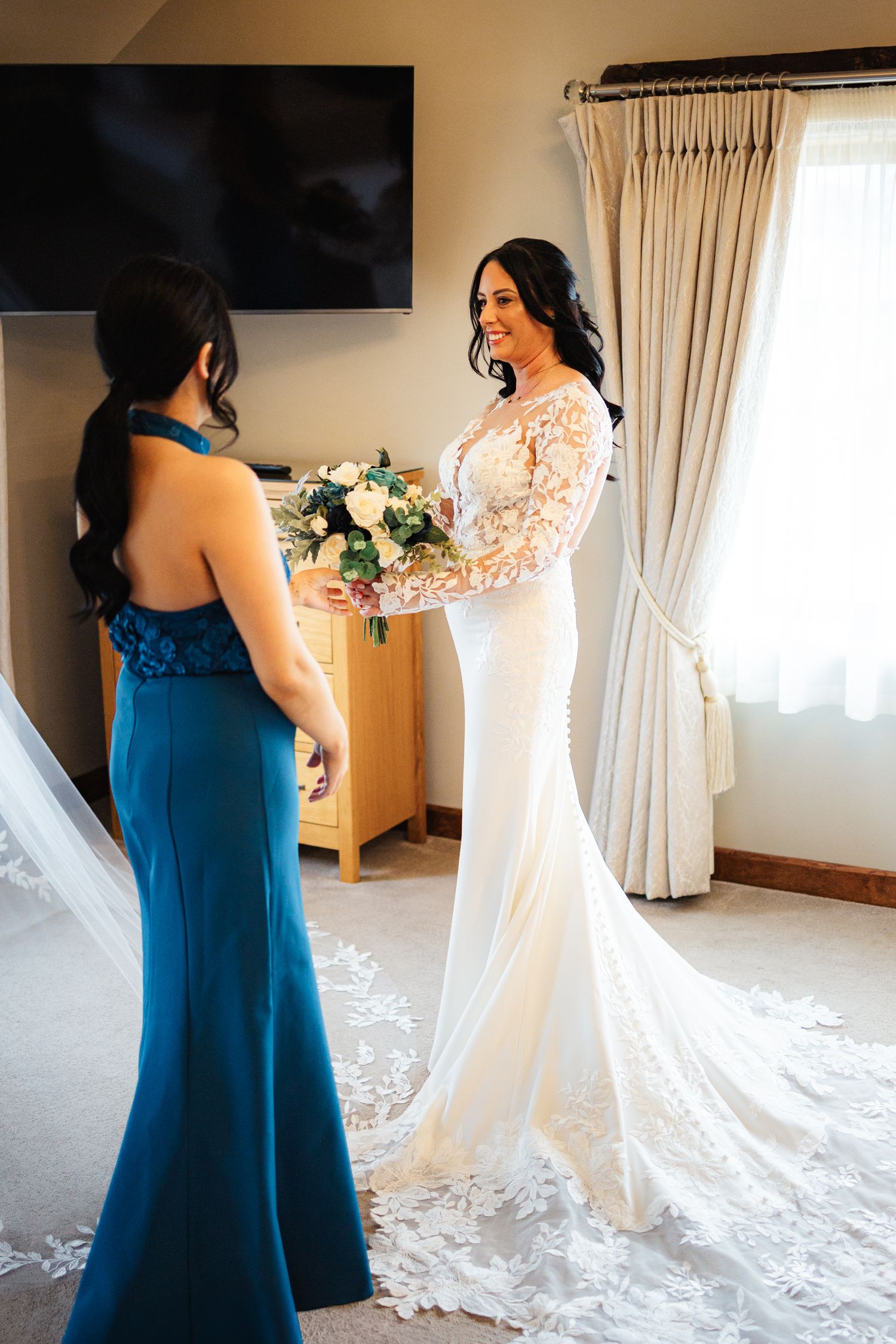 Bride in lace gown holds bouquet with bridesmaid in teal dress during wedding preparations in elegant room.