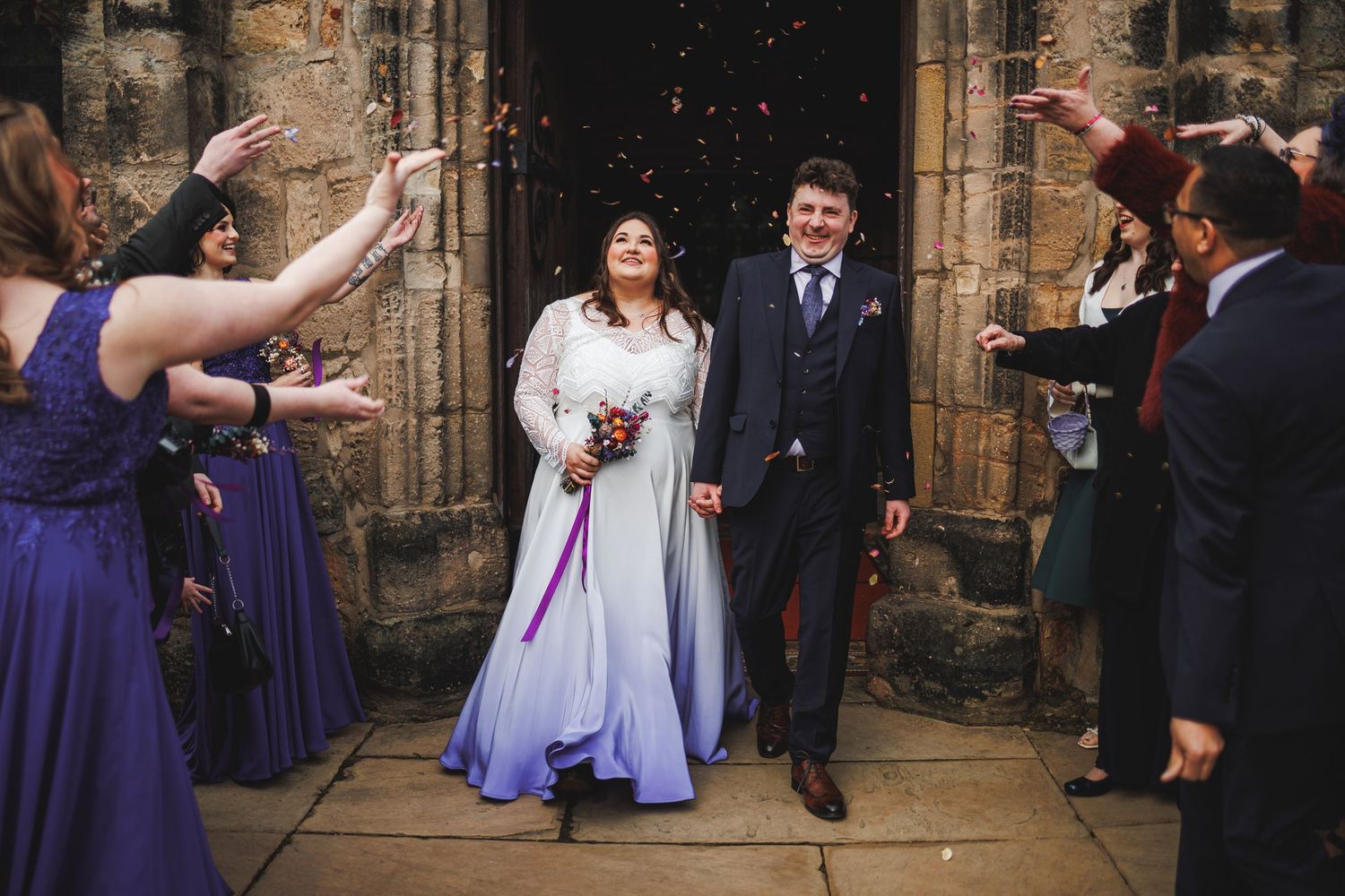 Bride in lavender gown and groom exit church as wedding guests throw confetti in joyful celebration.