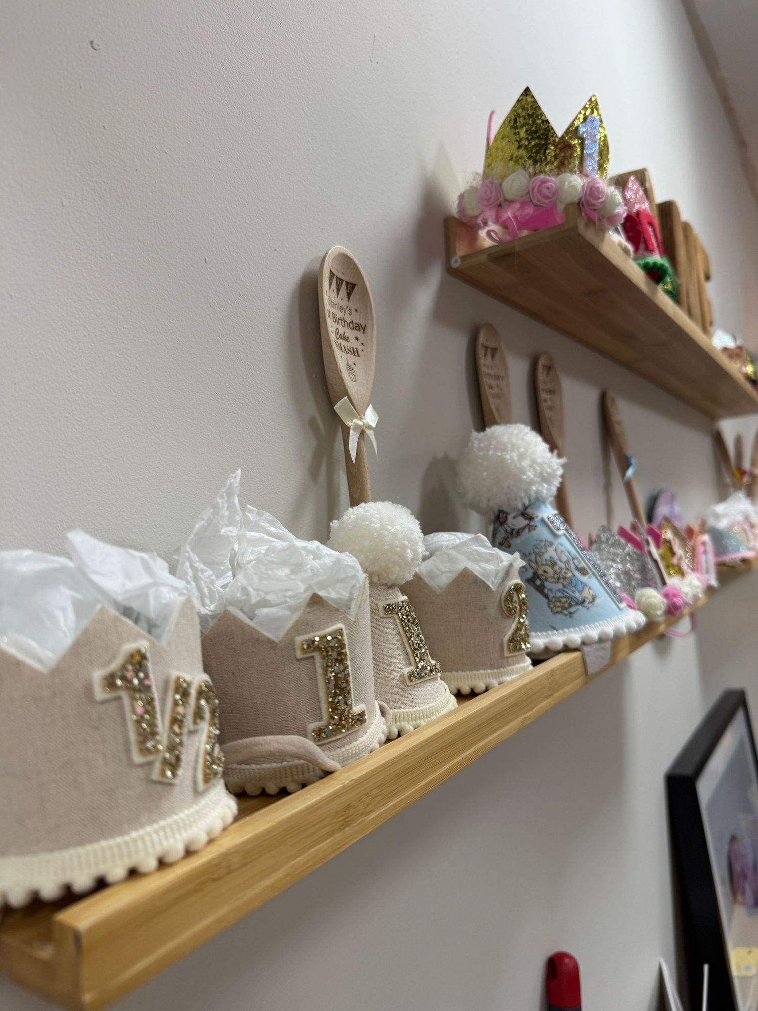 Decorative wooden shelves displaying handmade first birthday crowns with gold number one, pom-poms, and festive party hats.