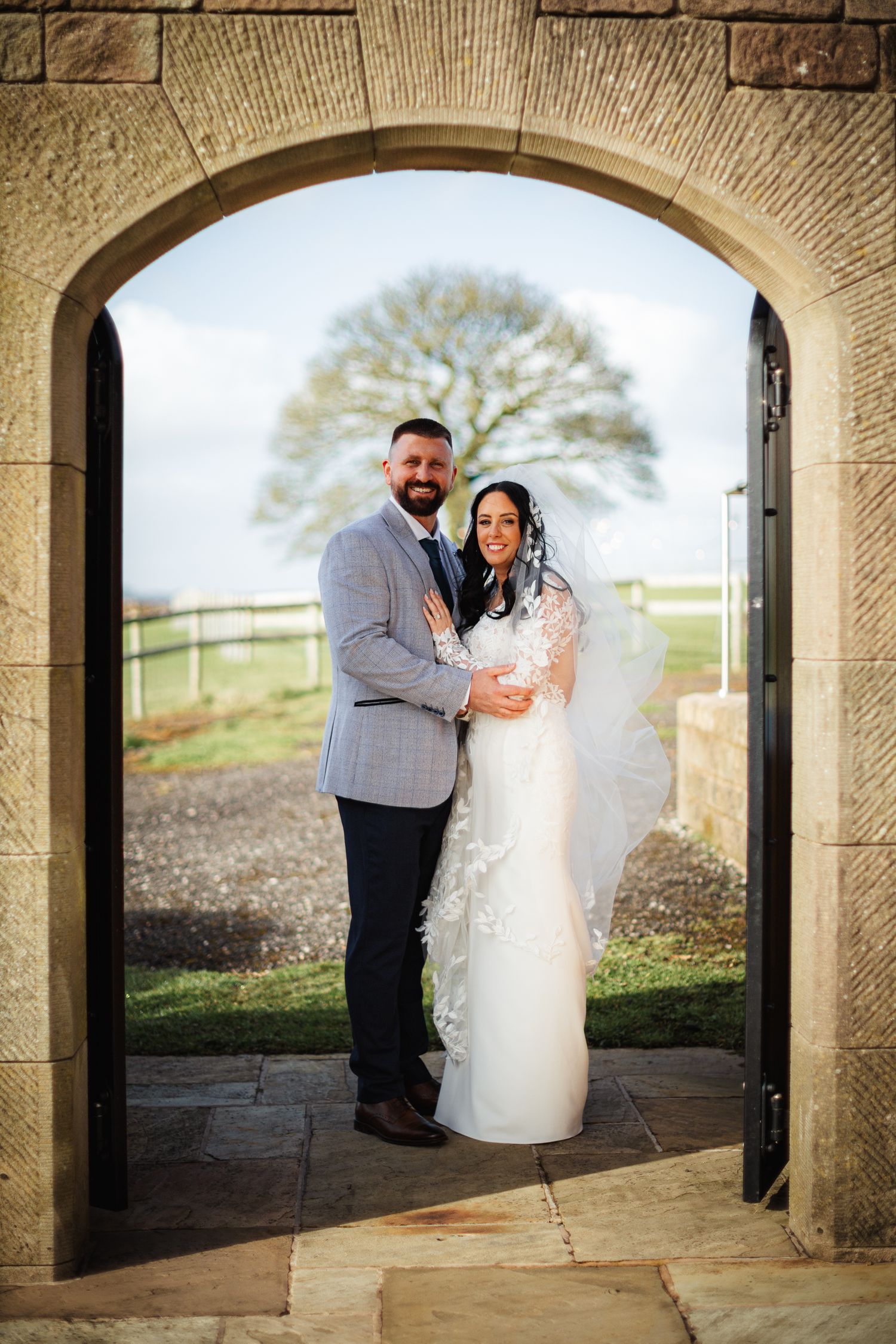 Bride and groom posing in a stone archway doorway at a countryside wedding venue, with a tree visible in the background.