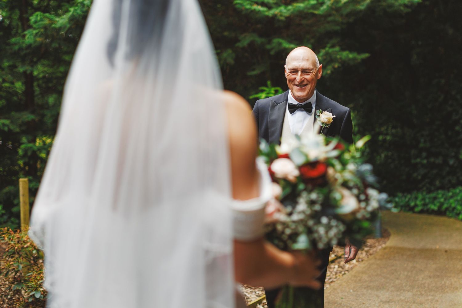 Father smiling joyfully at bride in white gown and veil holding floral bouquet during outdoor wedding ceremony.