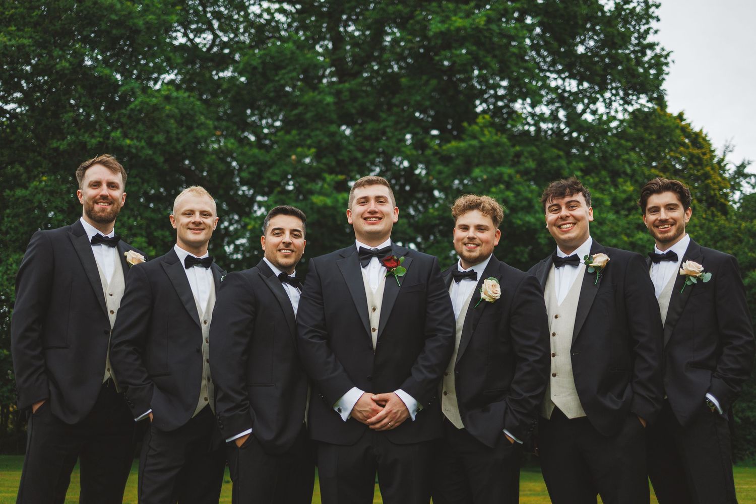 Seven groomsmen in black tuxedos and bow ties pose outdoors on green lawn with trees behind them.