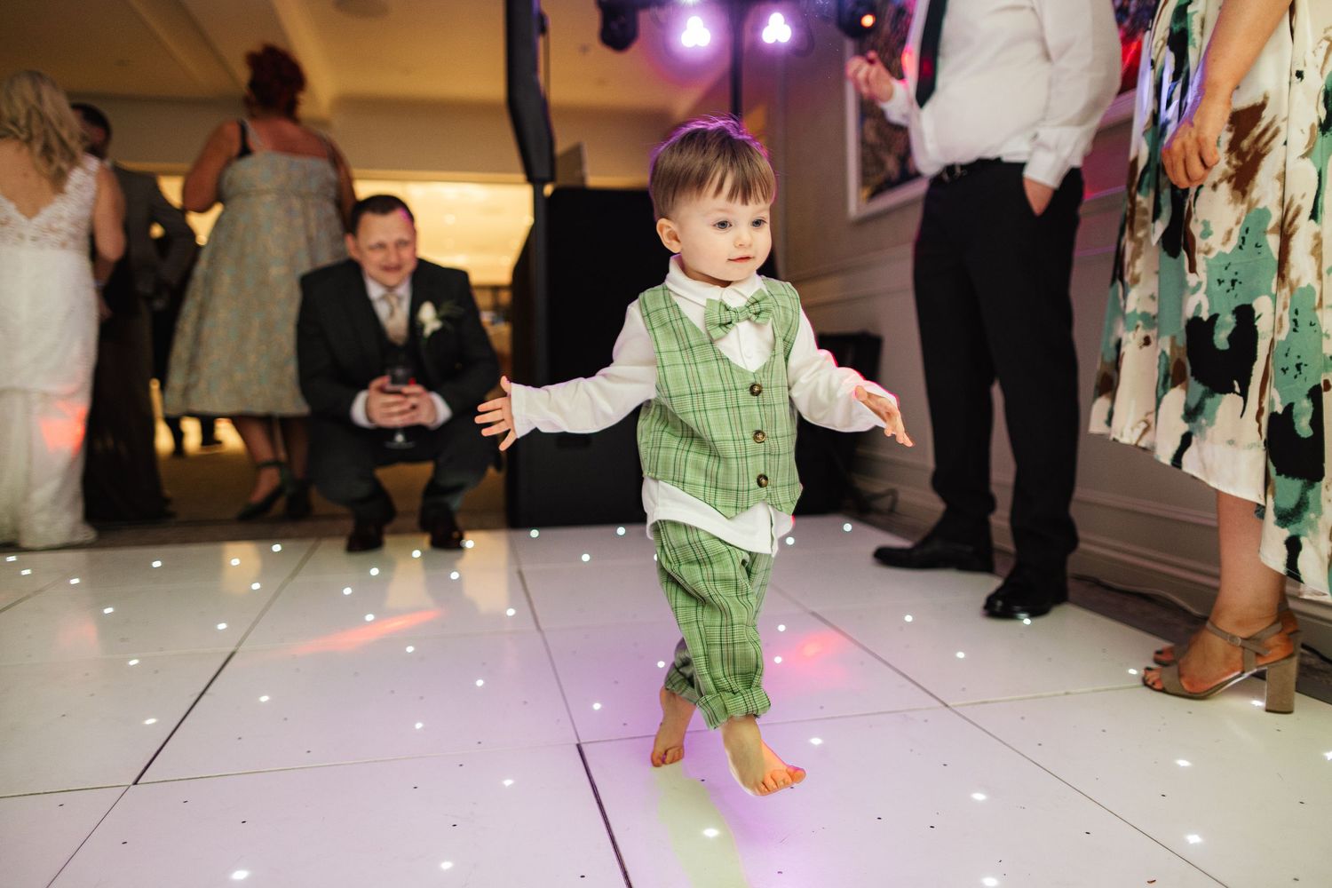 Adorable toddler boy in green plaid vest and bow tie dancing barefoot on a sparkly white LED dance floor at a wedding.
