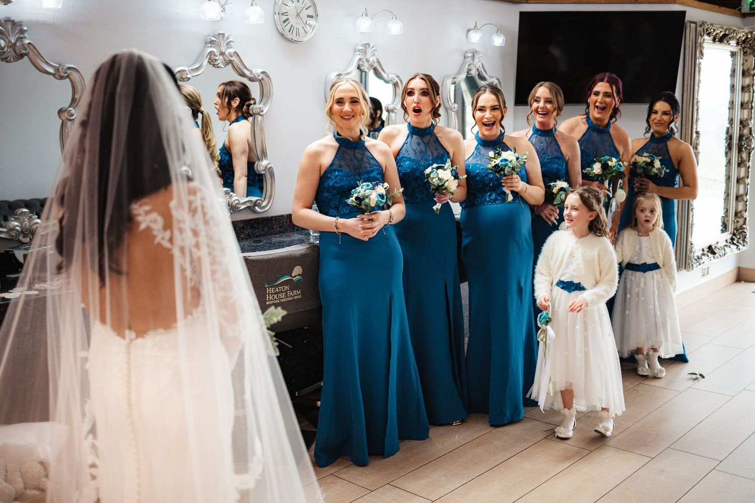 Bride in white gown and veil reveals herself to bridesmaids in teal dresses holding bouquets, with flower girl nearby.