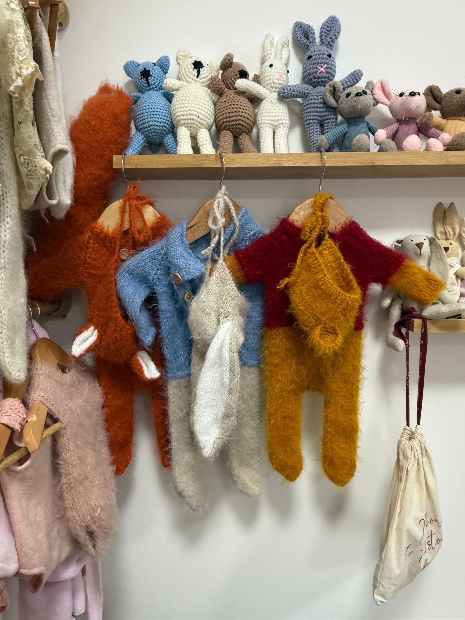 Handmade crochet animal toys on a wooden shelf with knitted baby clothing hanging on wall hooks below.
