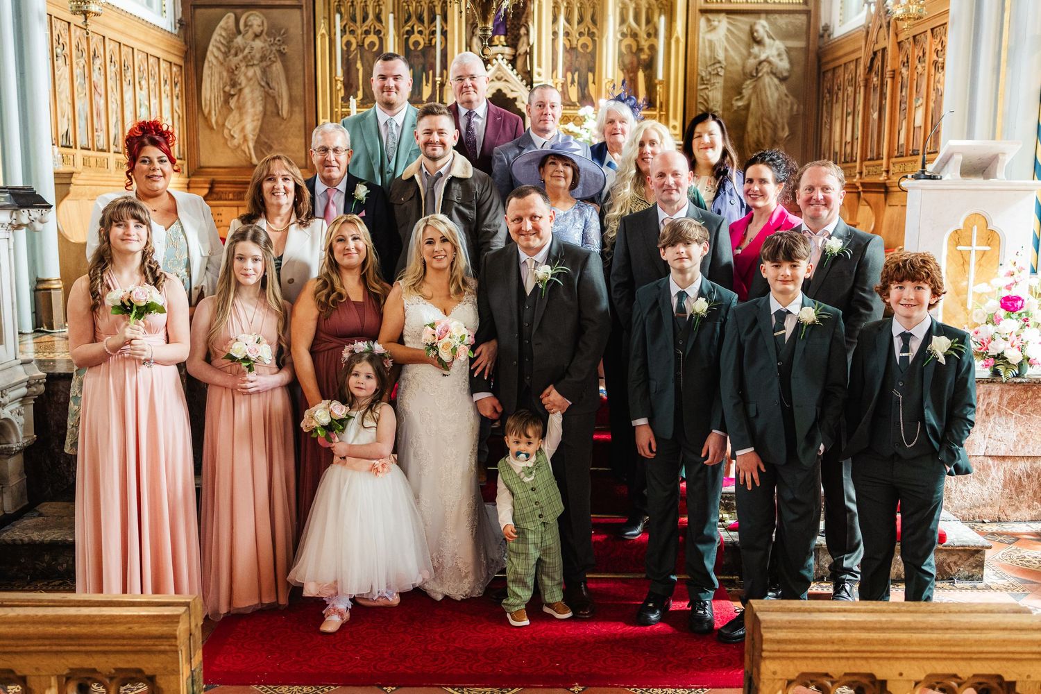 Wedding party group photo inside a church, featuring bride, groom, bridesmaids, groomsmen, and family on red carpet steps.