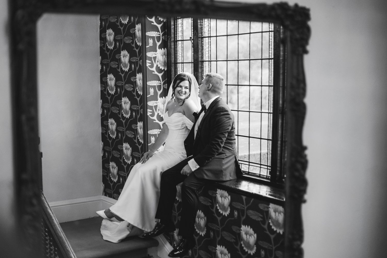 Black and white wedding photo of bride and groom sitting intimately by a window, framed by an ornate dark mirror.