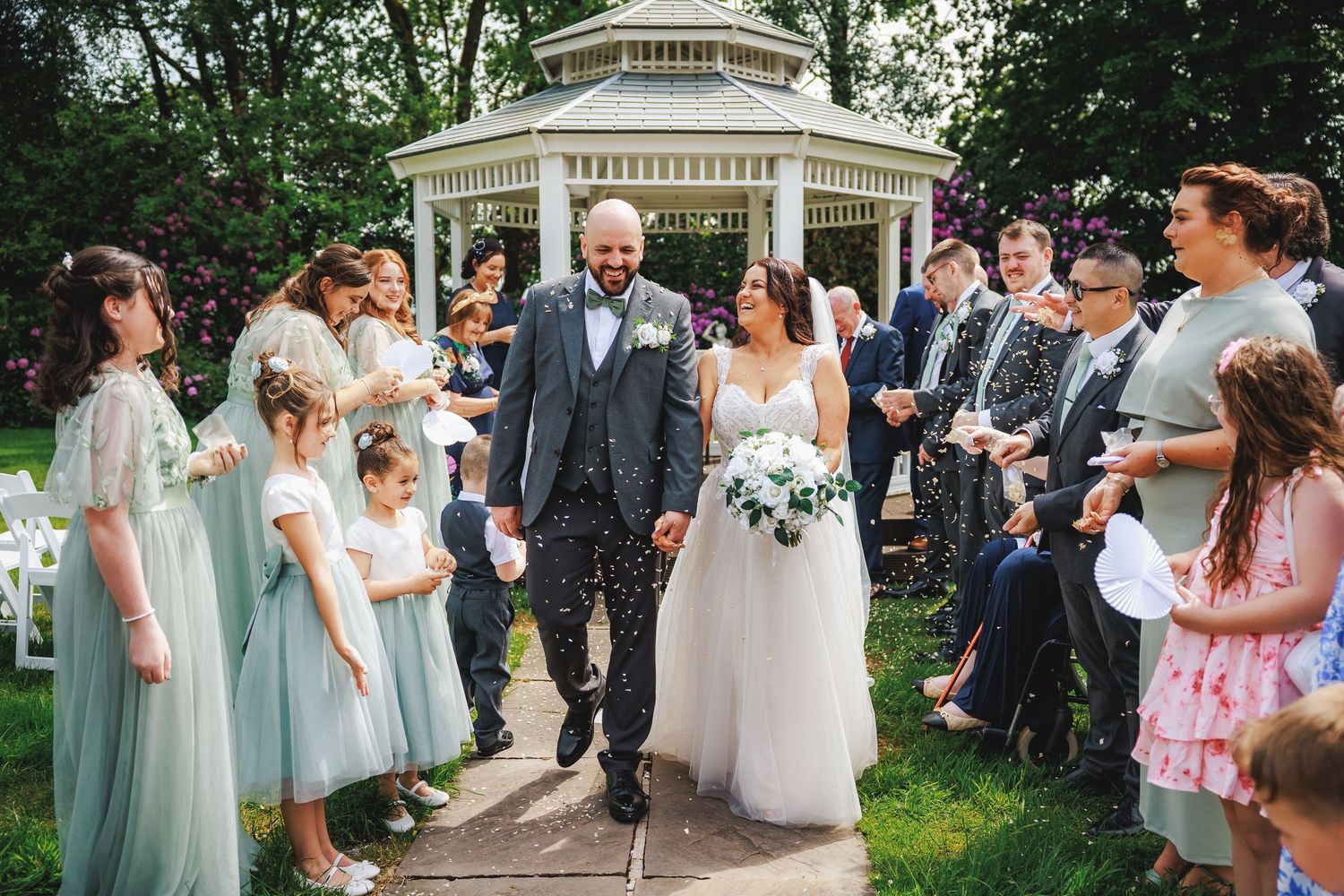 Bride and groom walk through confetti shower with wedding party outside a white gazebo in a lush garden.