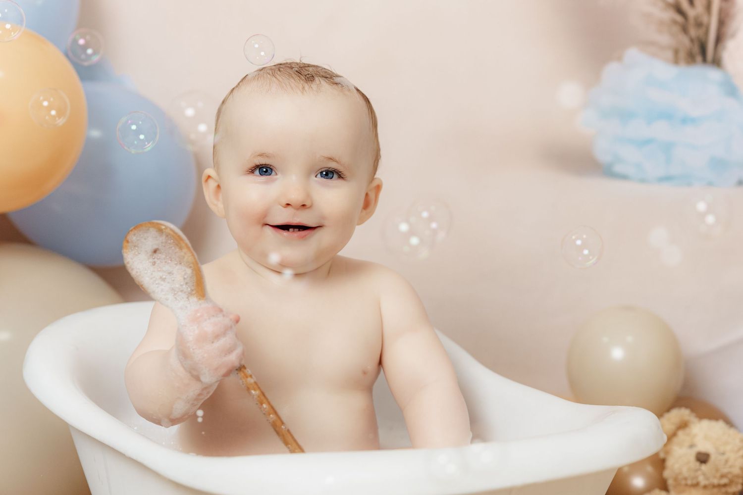Happy baby sitting in a white bathtub holding a wooden spoon, surrounded by colorful balloons and soap bubbles.