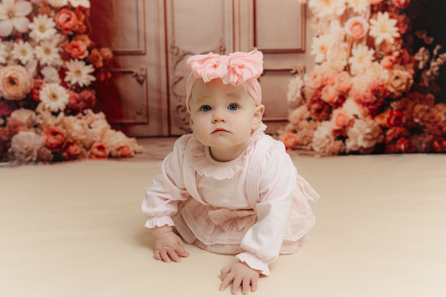 Baby girl in pink outfit and floral headband crawling on cream floor with flower backdrop.