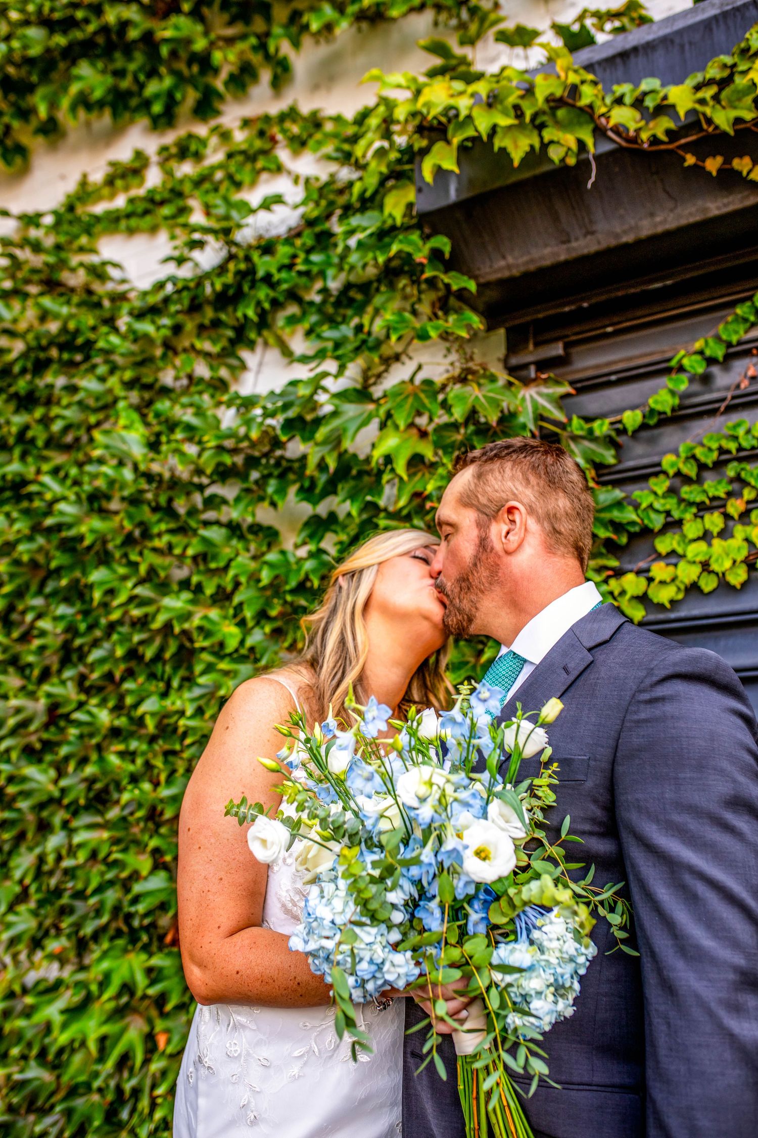 Wedding couple shares intimate kiss surrounded by lush green ivy backdrop.