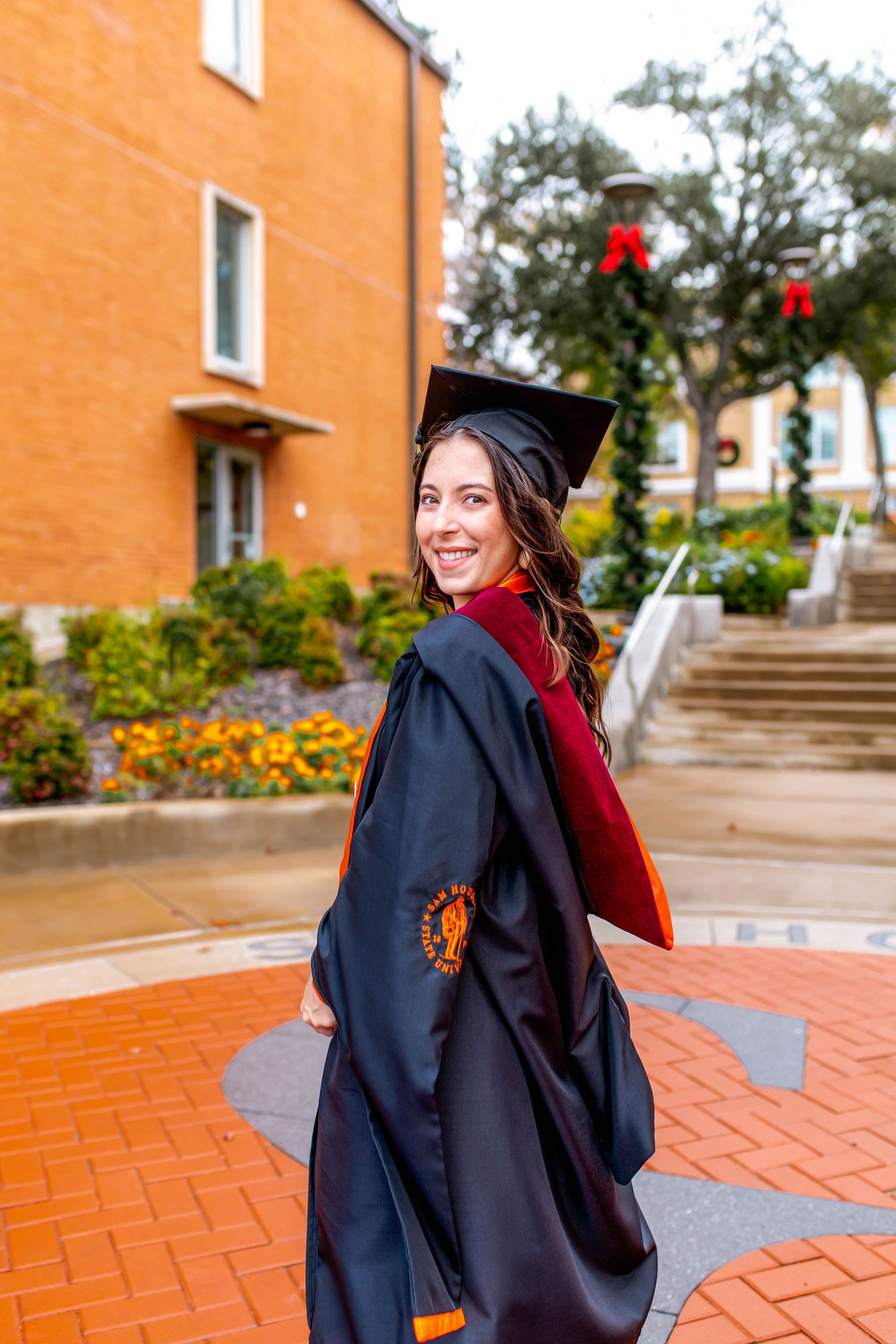 Two graduates in black caps and gowns embrace on red brick pavement during graduation.