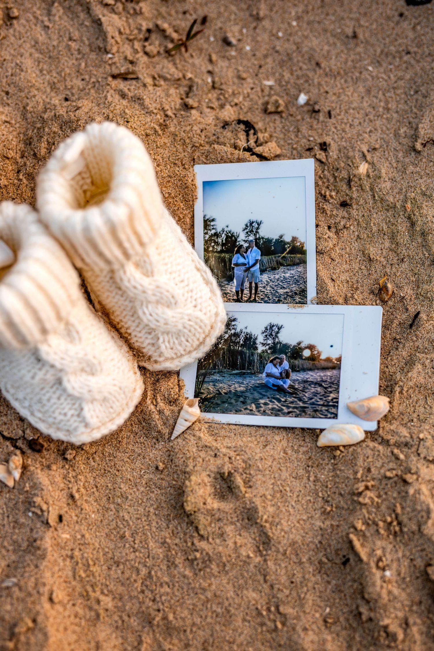 White knit baby booties placed next to polaroid photos on sandy ground in artistic arrangement.