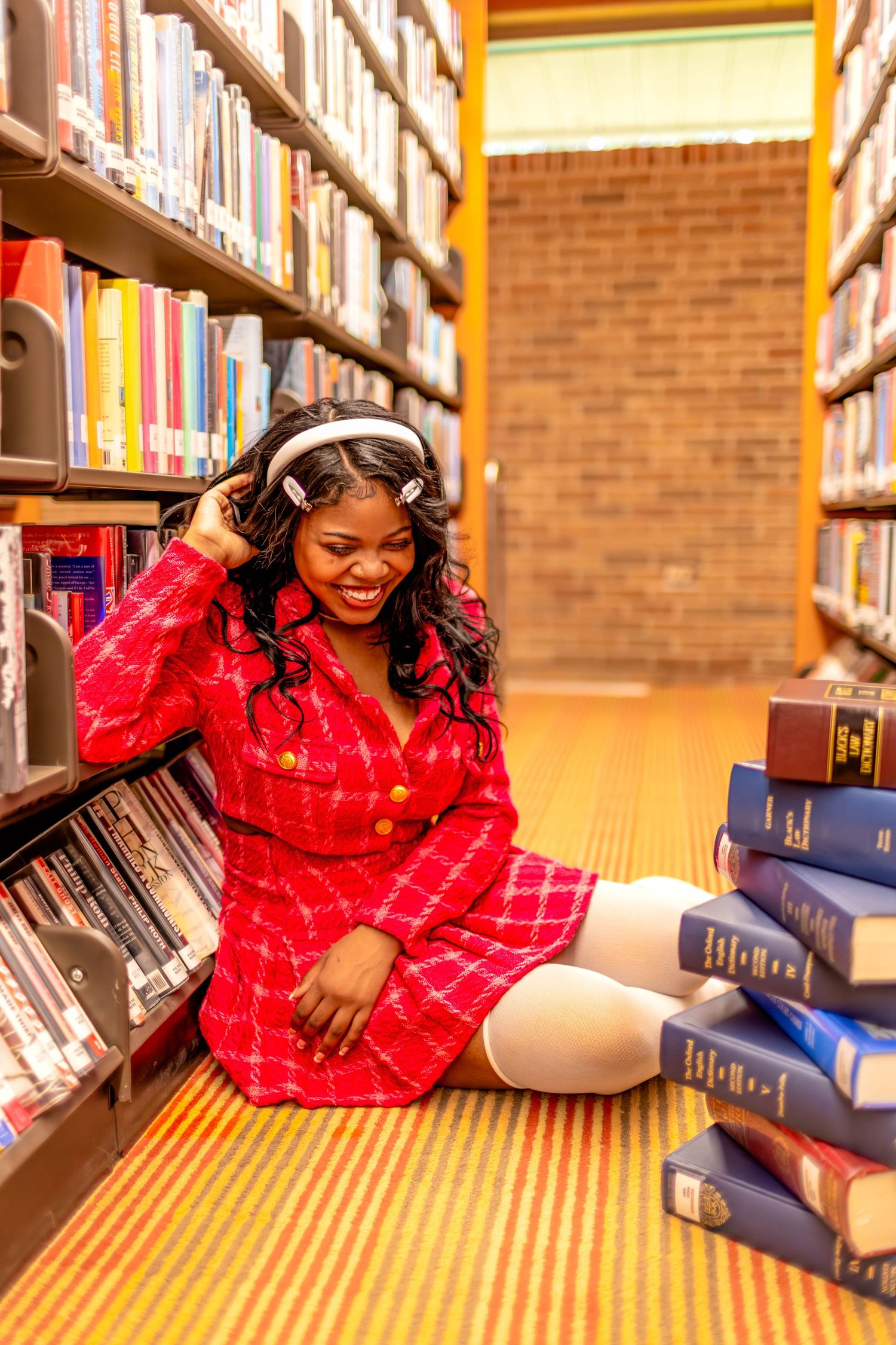 Joyful library photoshoot with books and red dress on wooden floor.