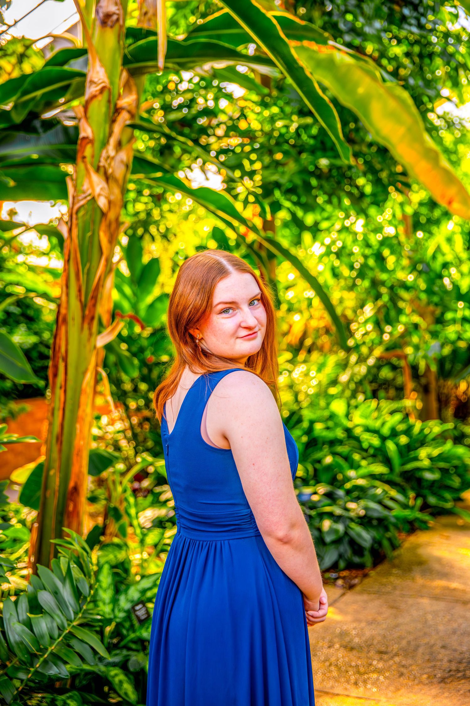 High School Senior in blue dress smiling during outdoor photo session in lush garden setting.