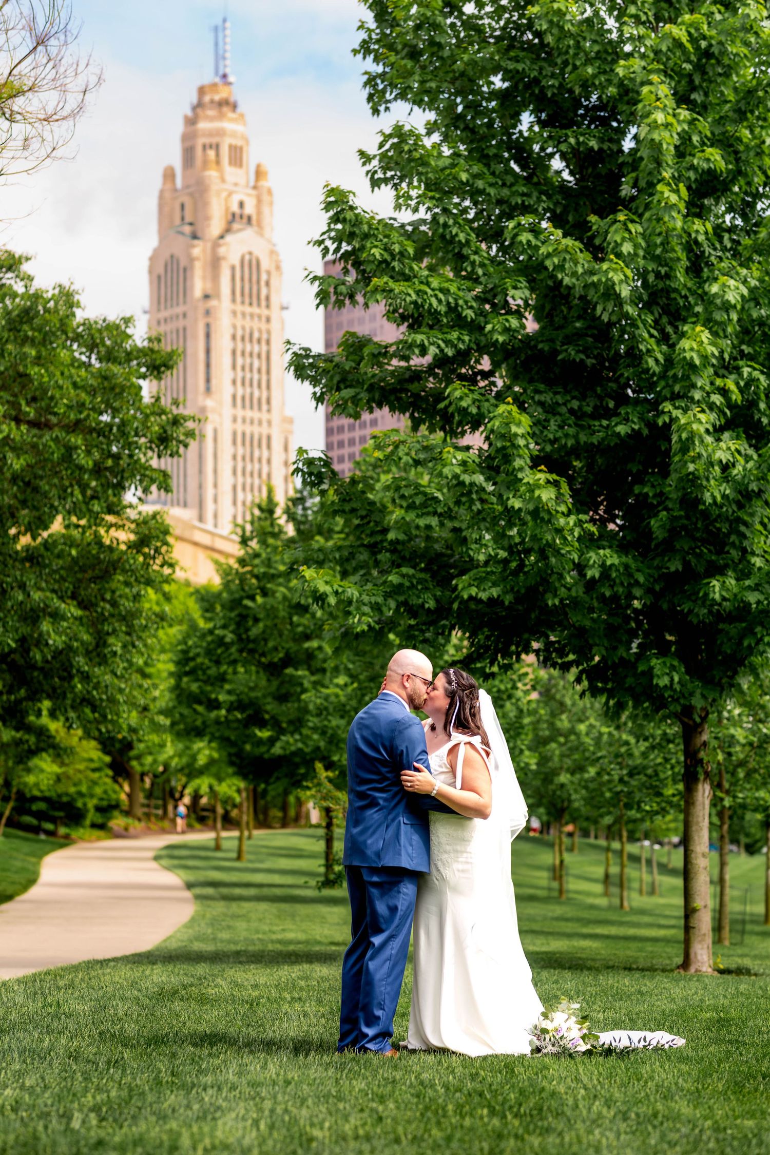 A romantic wedding scene in a lush green park with a towering historic building visible in the background.