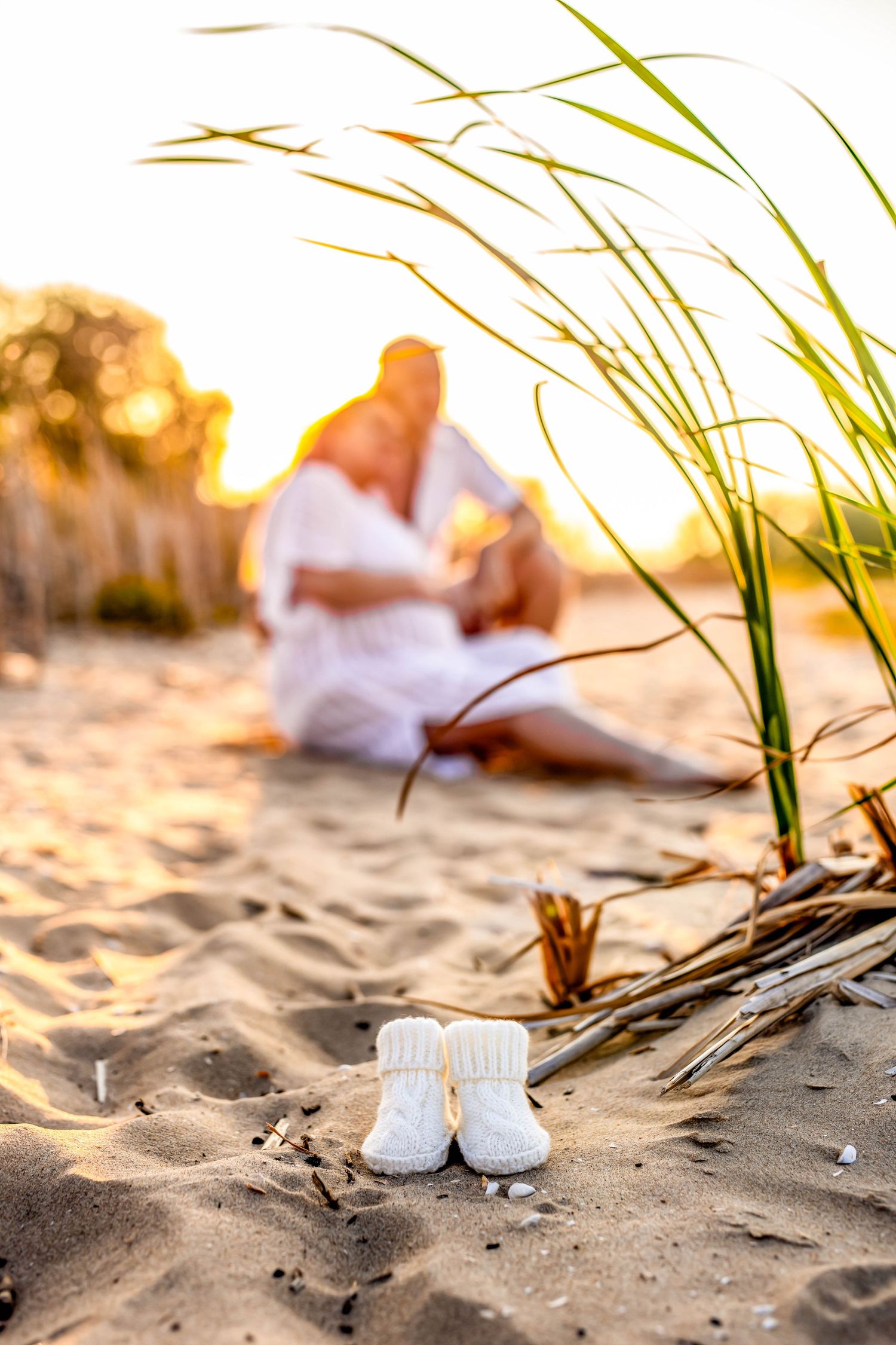 Small white baby shoes sit in the sand during golden hour on a beach with grass in the background.