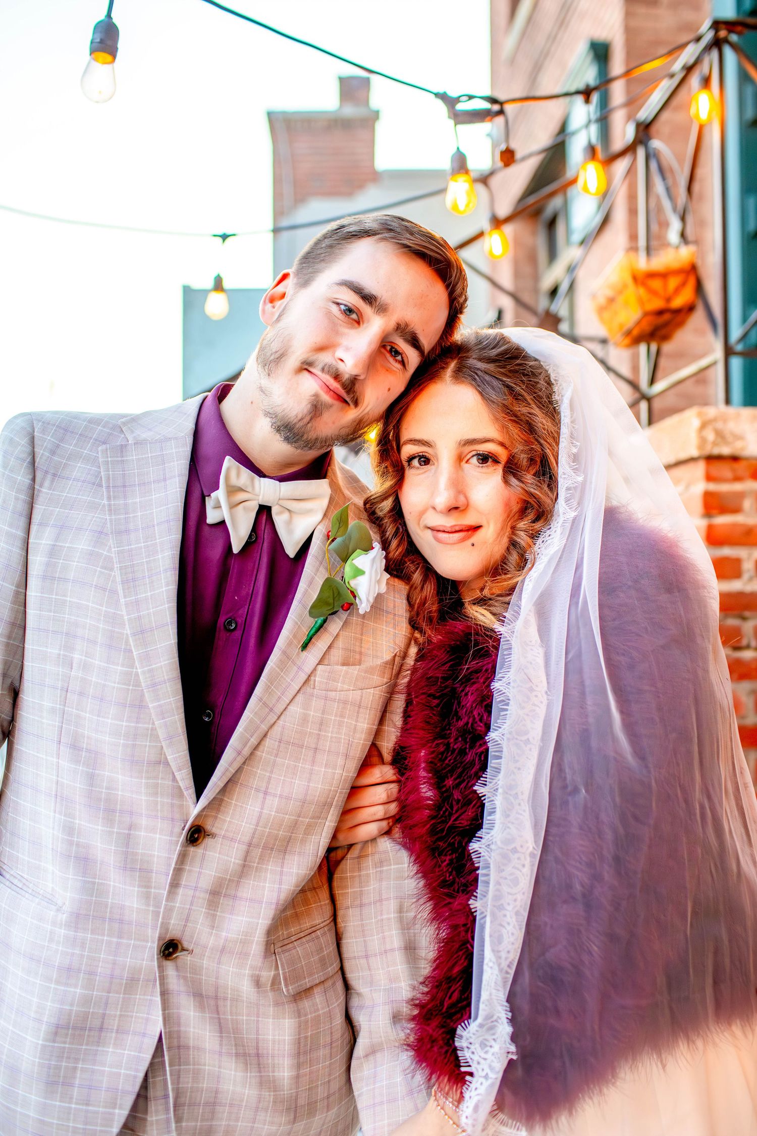 Evening wedding portrait against brick architecture with festive string lights overhead.