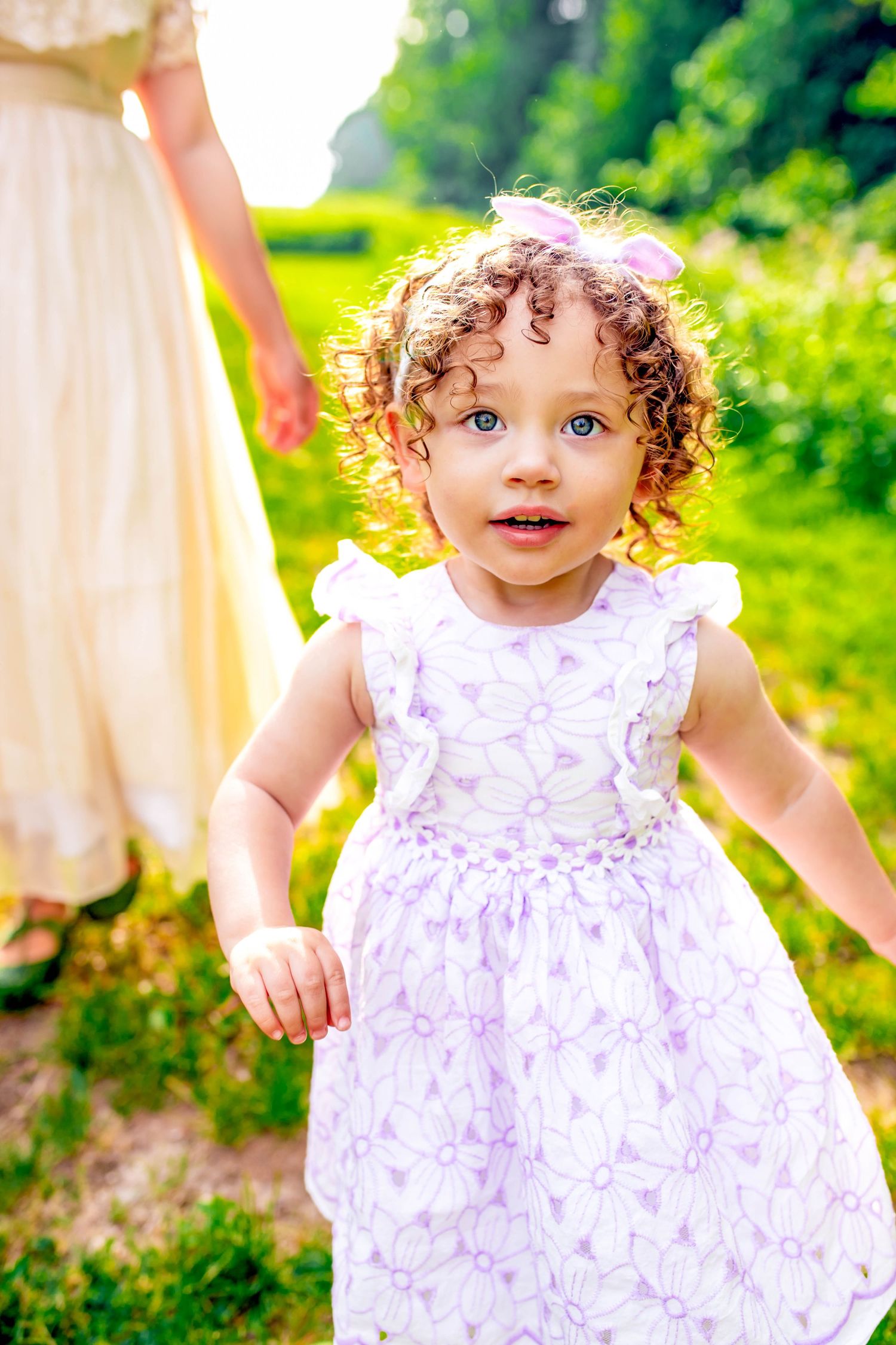 Toddler in white floral dress plays joyfully in a sunny garden setting.