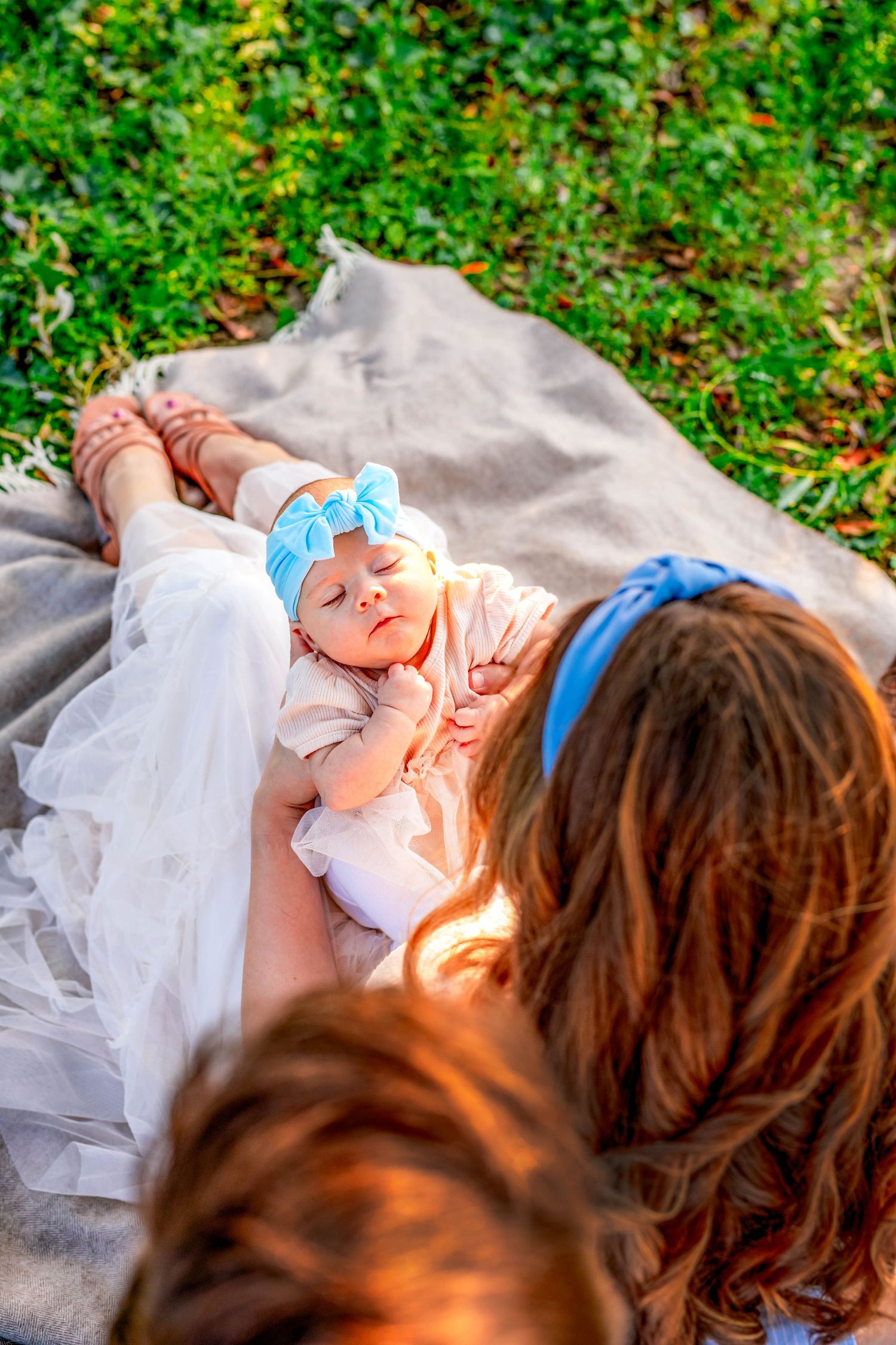 Newborn baby lying on soft blanket outdoors with greenery in background under gentle sunlight.