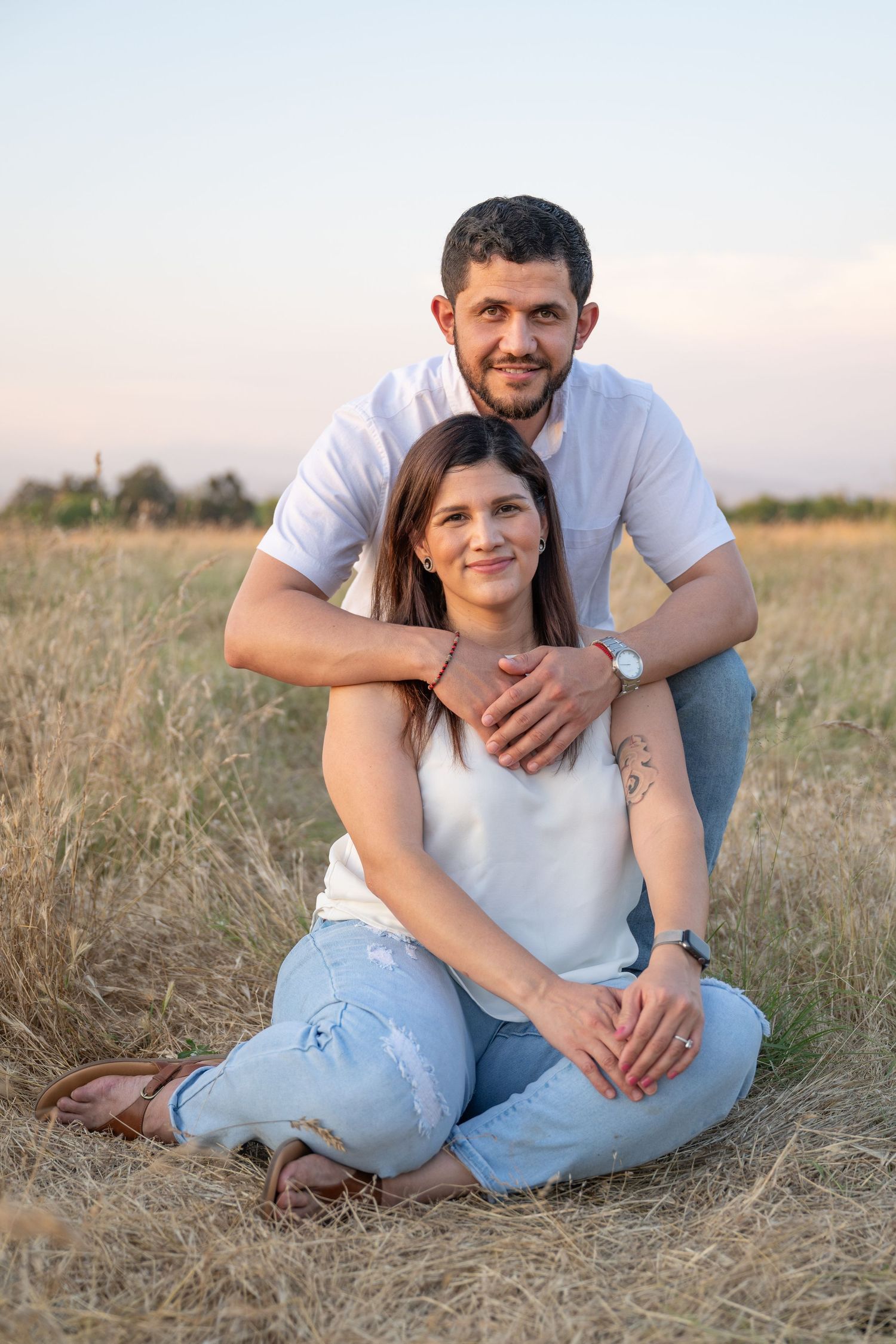 Couple poses together in casual wear on dried grass field during sunset.