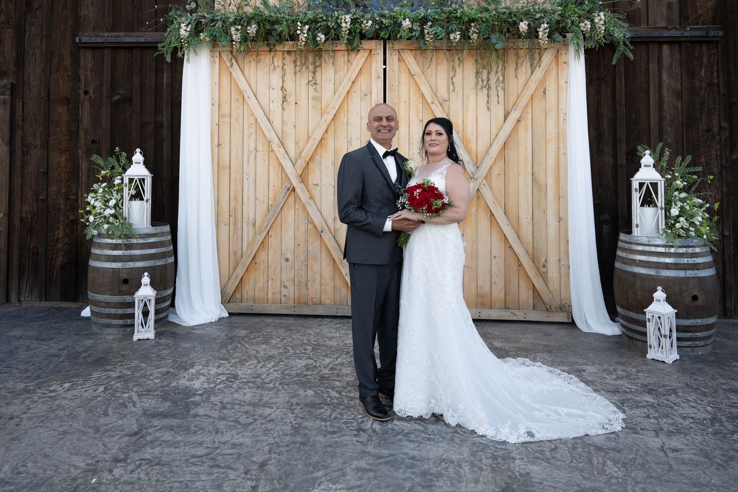 A couple poses in front of rustic barn doors decorated with greenery and white lanterns for their wedding day.