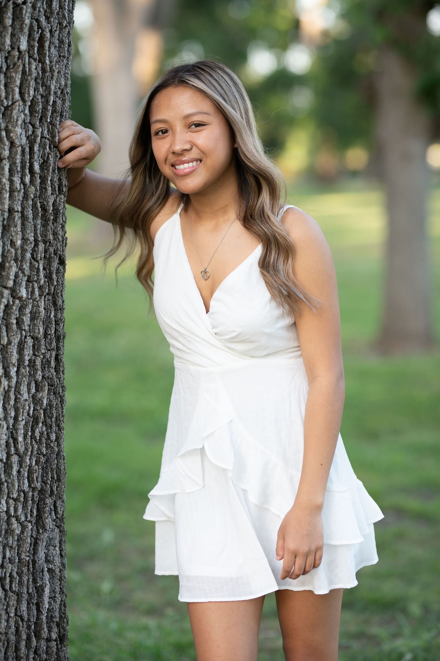 A person in a white ruffled romper poses outdoors against trees in a park setting during a summer photoshoot.