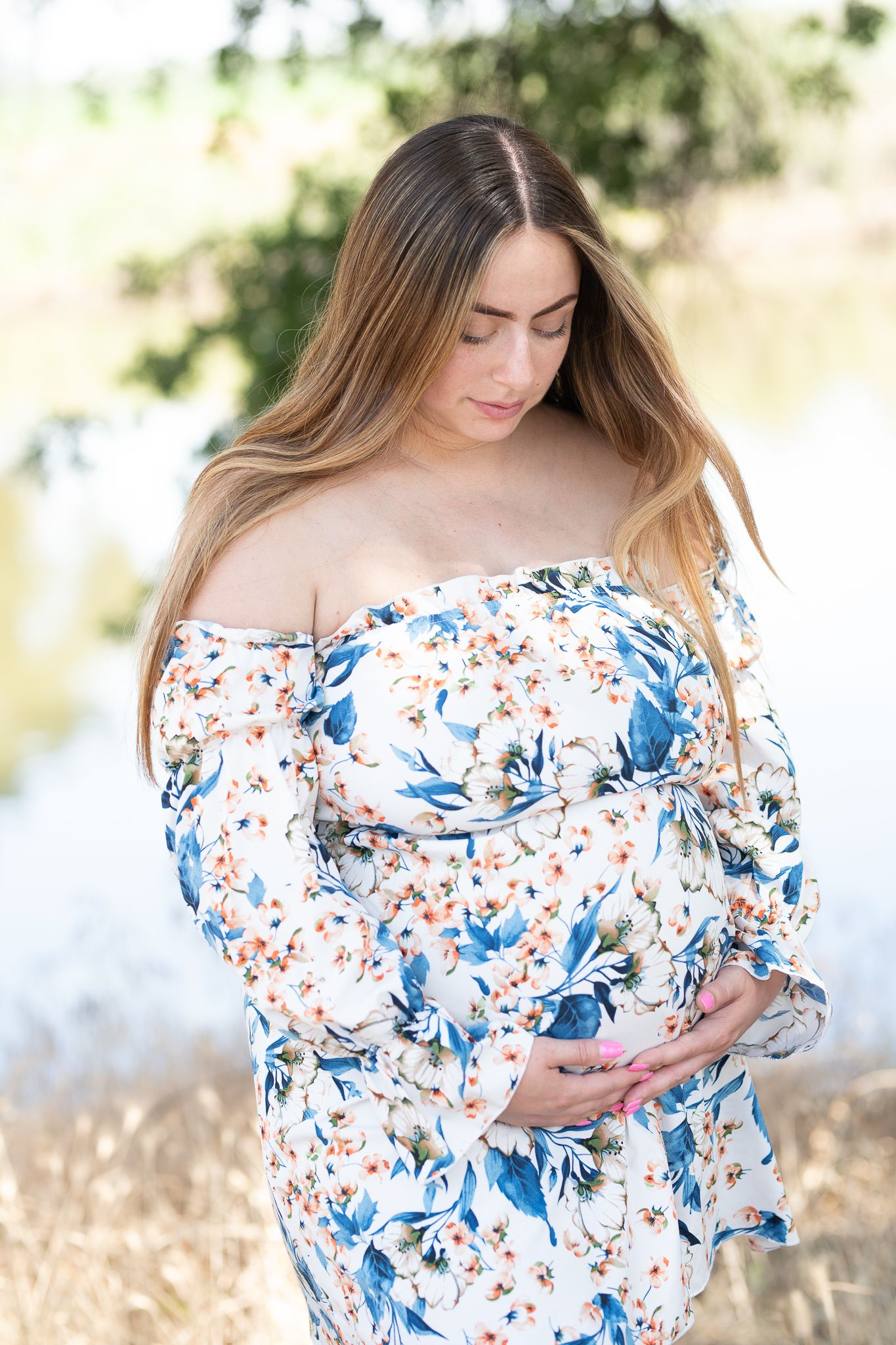 Person in dreamy outdoor setting wearing white off-shoulder dress with blue floral pattern in warm sunlight.