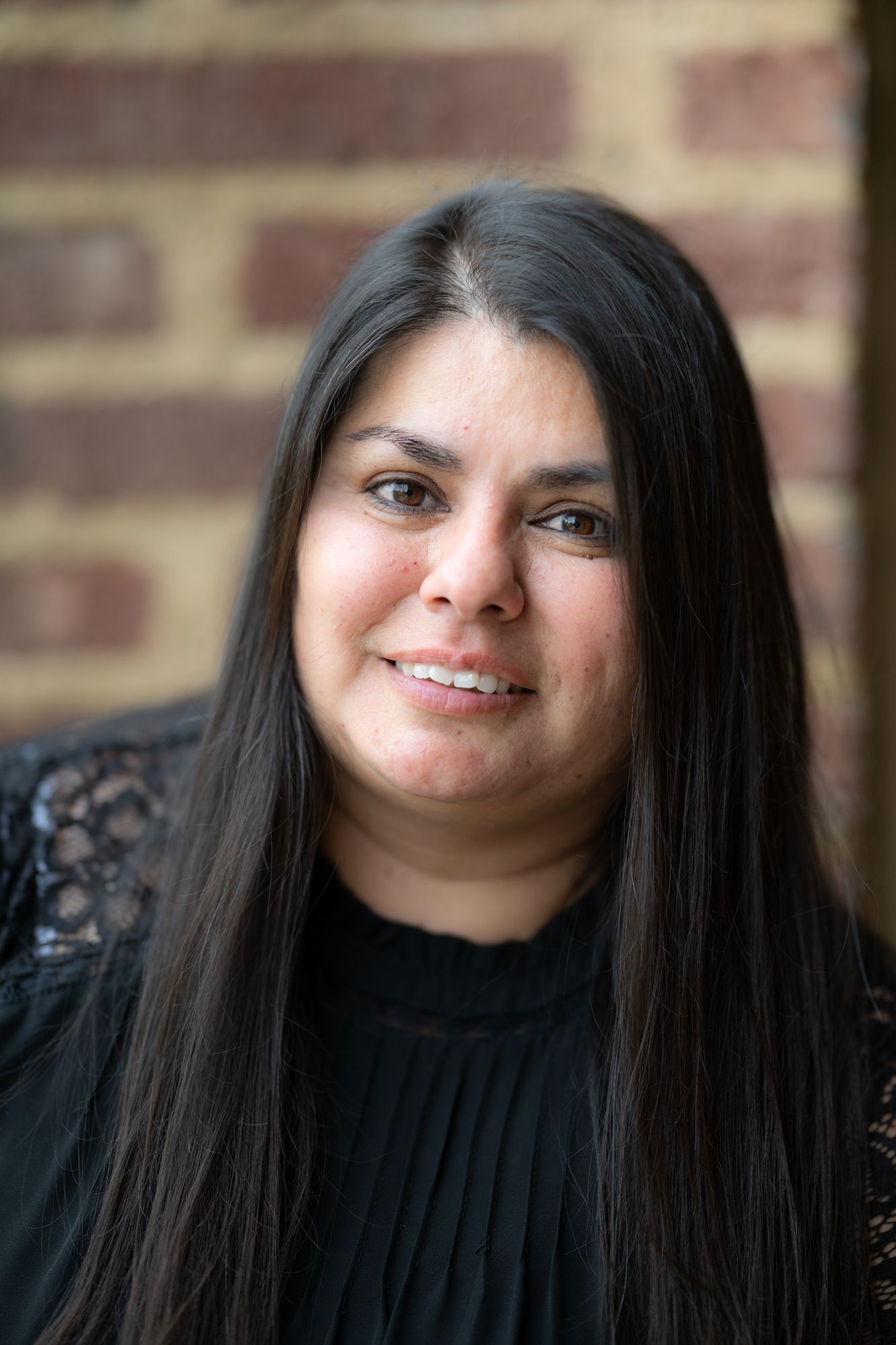 Portrait of someone with long dark hair wearing a black top against a brick wall.