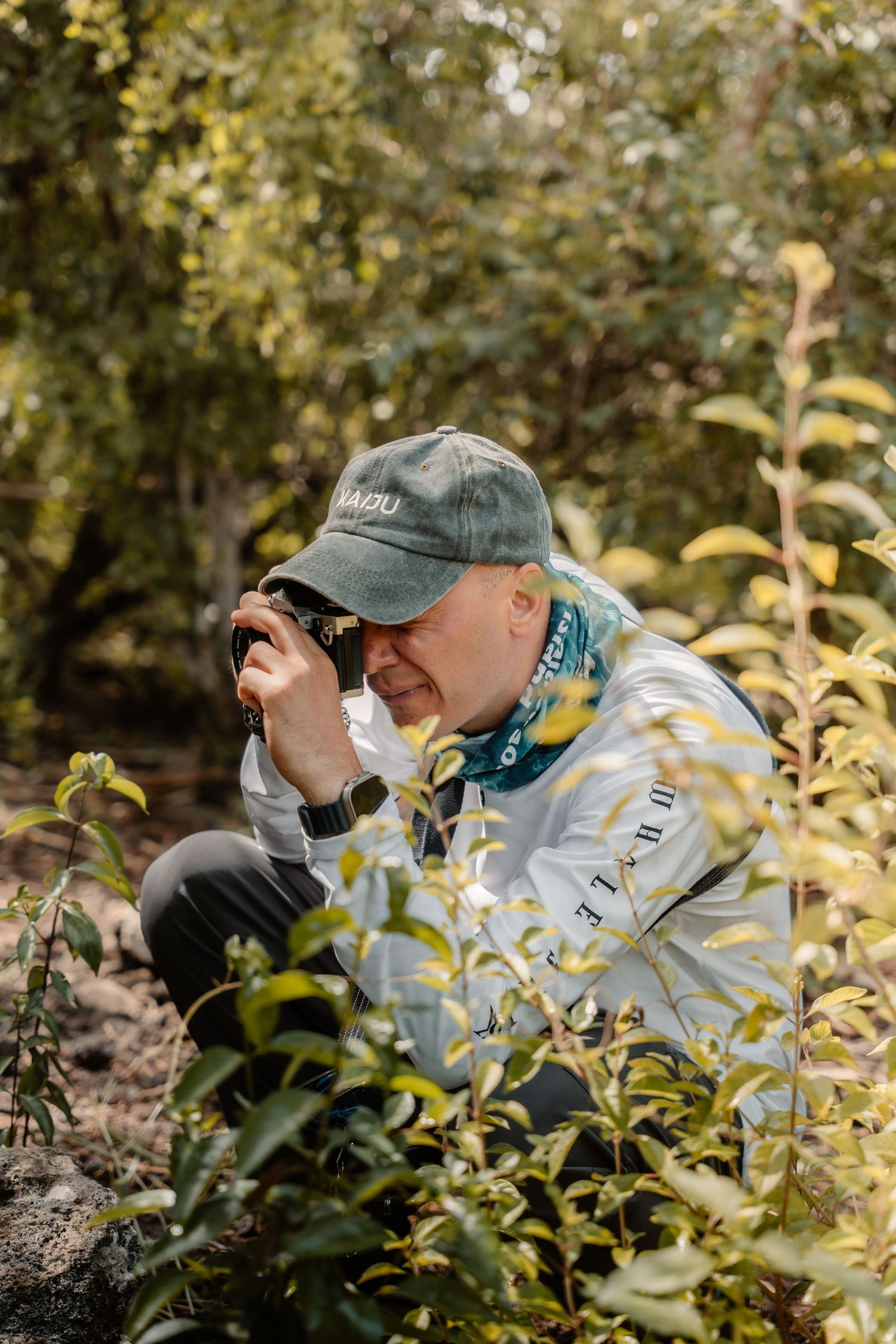 Man crouching in lush green forest, taking photos with camera, wearing green cap and light jacket.