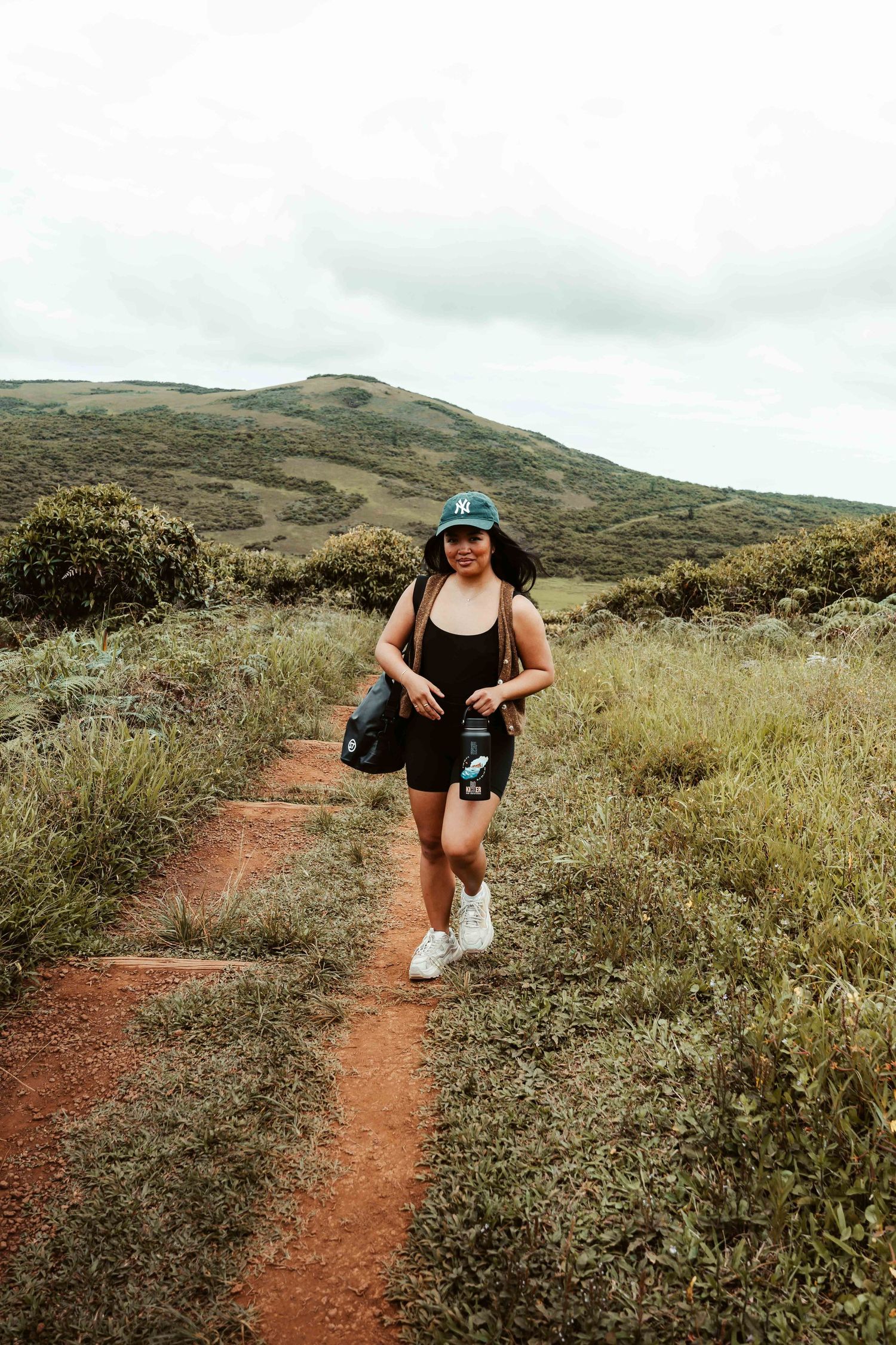 A hiker in athletic wear walks along a scenic dirt trail winding through green hills under cloudy skies.