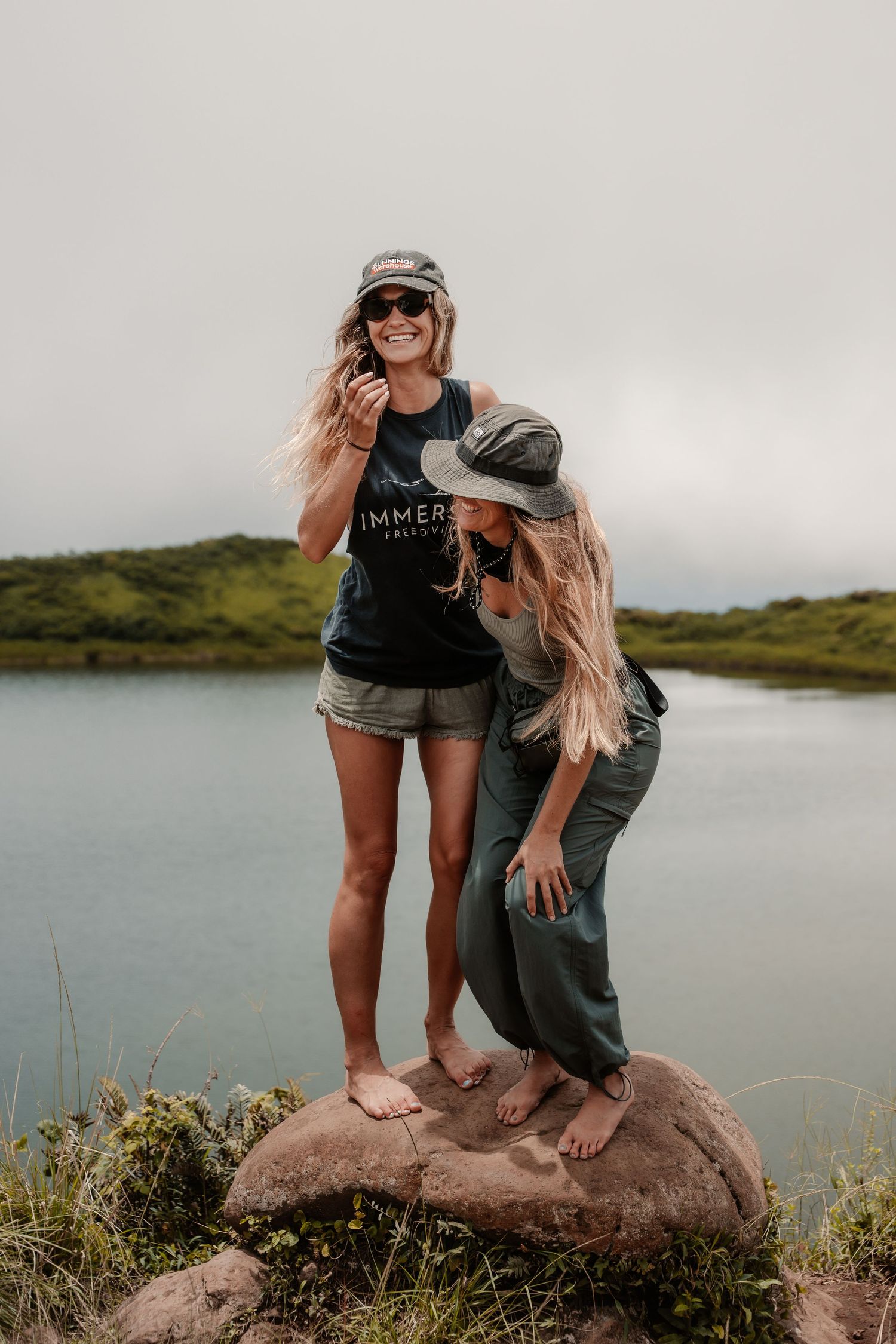 Two blonde women standing on a rock by a scenic lake, one wearing a black tank top and sunglasses, both in hats.
